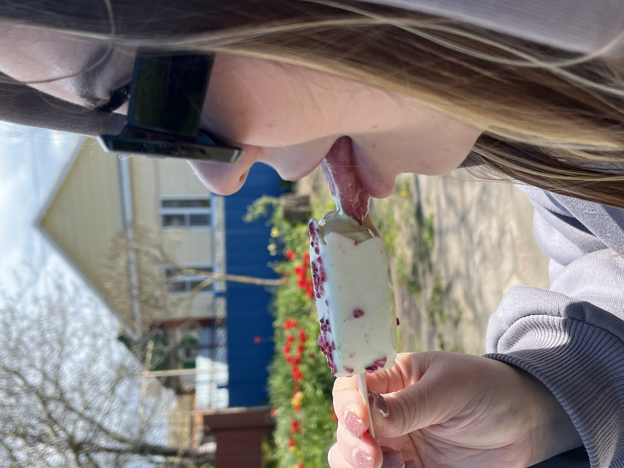 A woman with blonde hair is eating a white ice cream bar with red sprinkles. She is wearing a baseball cap and black sunglasses. The background features a house with yellow and blue siding, along with red tulips and trees. The location is outdoors, and the image is taken in Port-Katon, Russia.