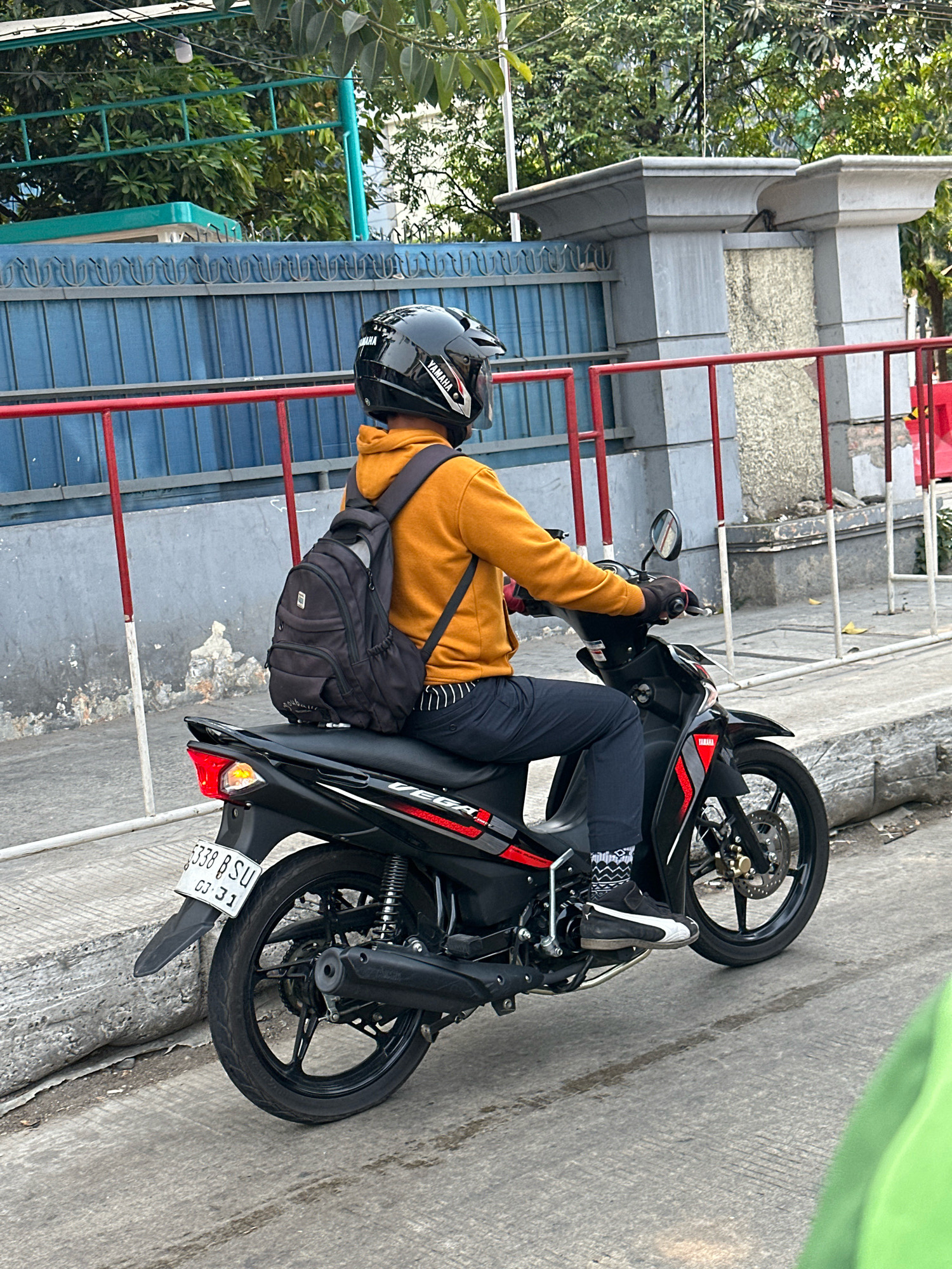 A person wearing a yellow hoodie, black helmet, and dark pants is riding a black Yamaha motorcycle. The person has a black backpack on their back and is wearing black gloves and patterned socks with black and white sneakers. The motorcycle has a license plate that reads "3338 B SU 03 31". The scene is in West Jakarta, Indonesia, as indicated by the street signs and architecture. The background includes a blue fence, a concrete wall, and trees. The lighting suggests it is daytime.