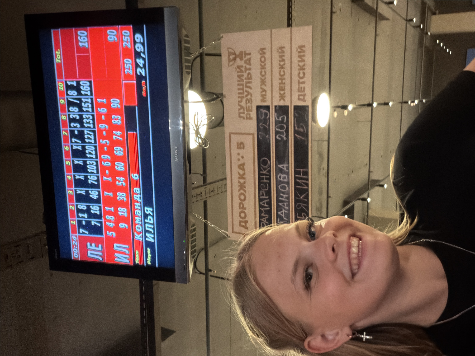 The media file captures a young woman with blonde hair, smiling and looking towards the camera. She is wearing a black top and a silver necklace with a cross pendant, and a silver cross earring. The background features a bowling score display on a television screen, showing scores and player names in Cyrillic. The display indicates a bowling game with scores for "ИЛЬЯ" (Ilya) and "ЛЕ" (LE) and "ИЛ" (IL), along with frame-by-frame results and total scores. To the right of the screen, a sign with Cyrillic text lists "ЛУЧШИЙ РЕЗУЛЬТАТ" (Best Result) and scores for "МУЖСКОЙ" (Men's), "ЖЕНСКИЙ" (Women's), and "ДЕТСКИЙ" (Children's) categories. The setting appears to be a bowling alley, with some lighting fixtures visible on the wall and ceiling. The overall mood is cheerful and relaxed, suggested by the woman's smile. The time displayed on the screen is "00:24", but this likely refers to the game time rather than the time of day.