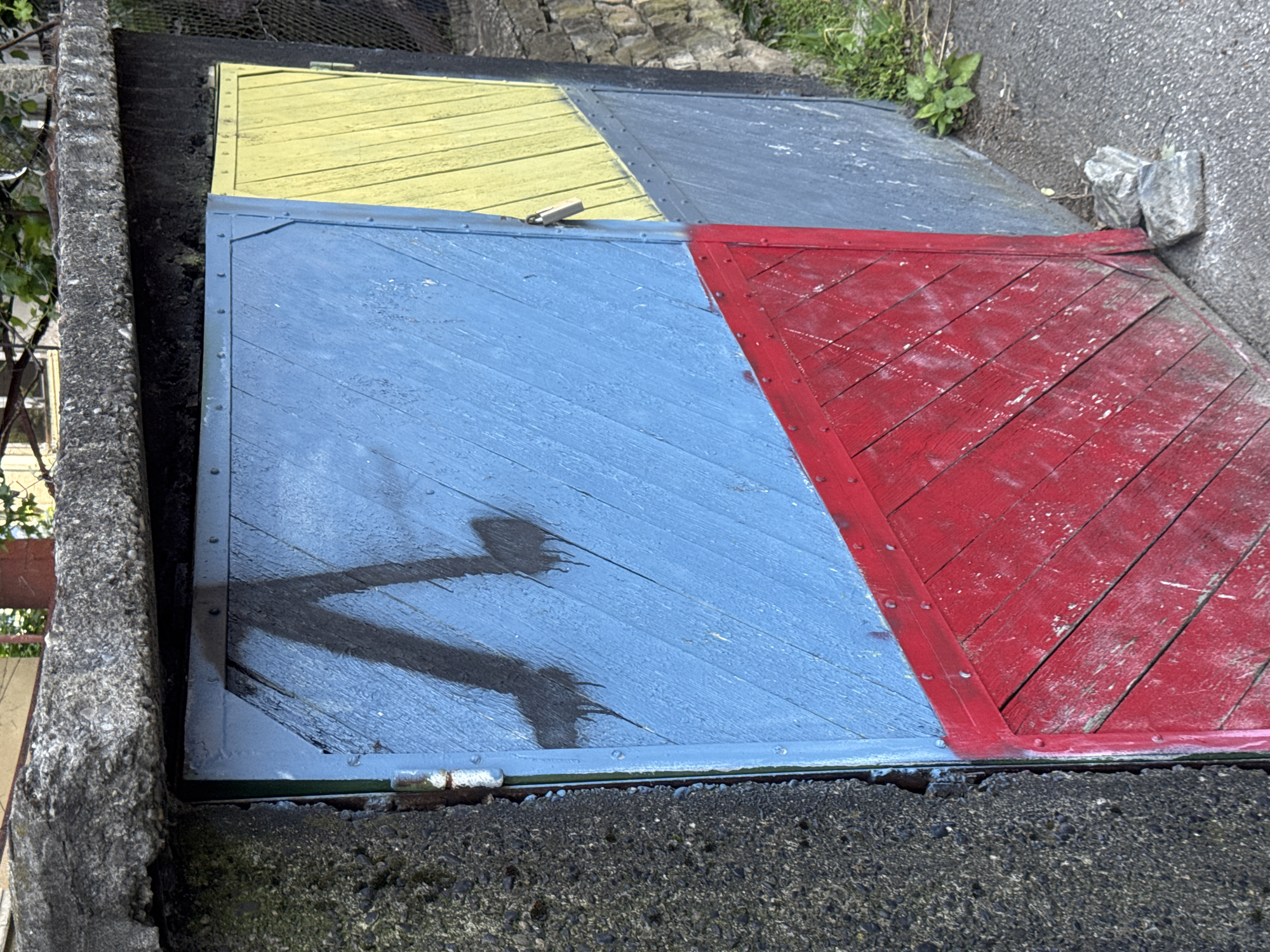 The image depicts a painted wooden cover embedded in a concrete structure in Lovech, Bulgaria. The cover consists of four colored sections: yellow, gray, blue, and red. The blue section has a shadow in the shape of a "V" on its surface. The cover is framed with metal and is bolted to the surrounding structure. The concrete structure is heavily textured and possibly weathered. Vegetation is visible near the cover.