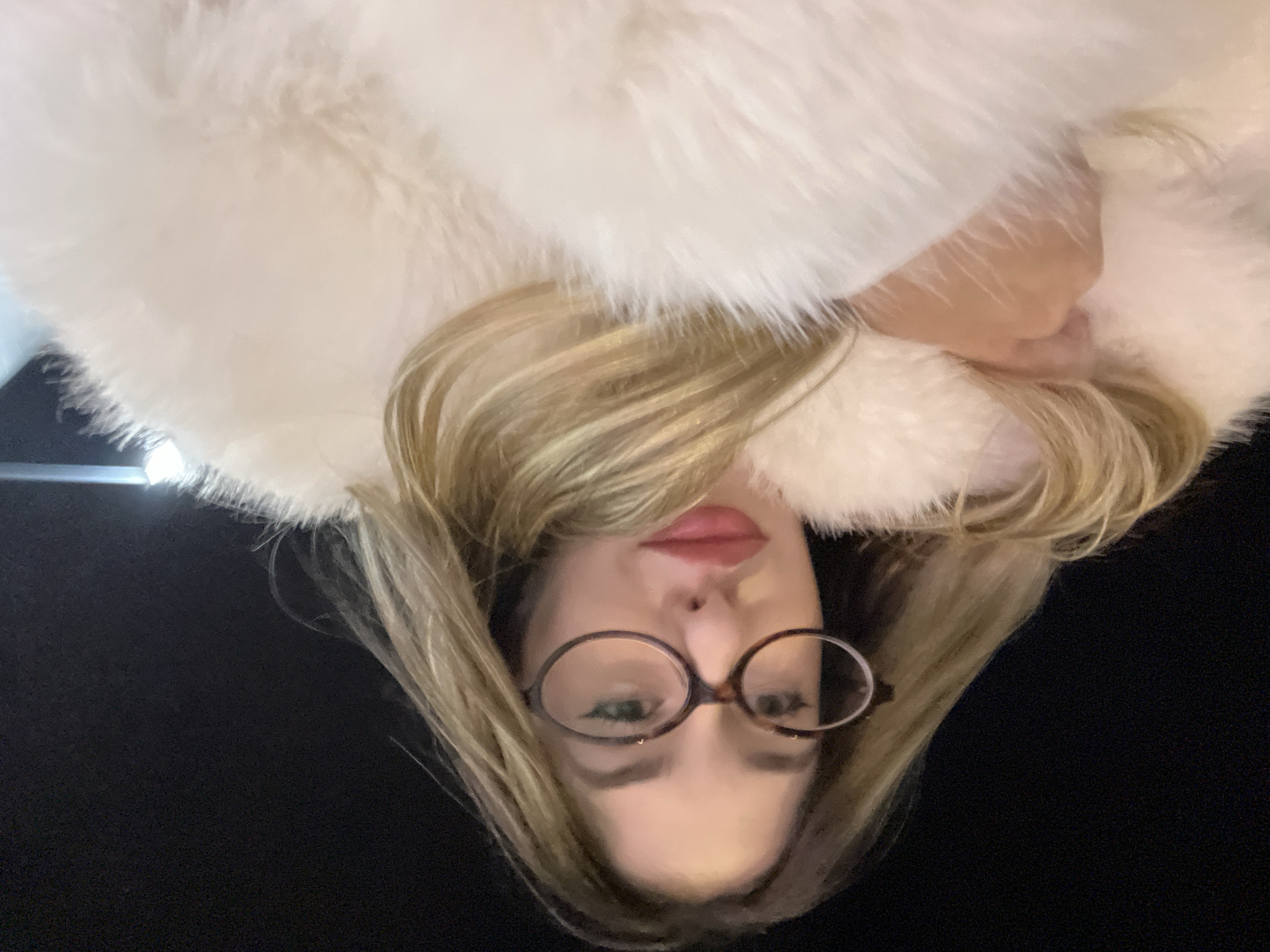 A woman with blonde hair and round glasses is facing the camera. She is wearing a white fur coat. The background is black. There is no evidence to indicate a location in Sofia, Bulgaria.