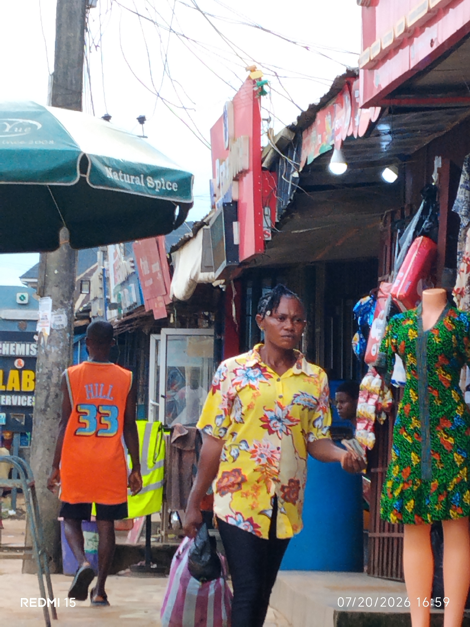 In Umuode, Nigeria, a street scene unfolds with a woman in a yellow floral shirt walking past a market stall. She carries a black bag in her right hand, and her left hand appears to be holding money or cards. To her left, a young man walks away from the camera, wearing an orange sleeveless shirt with "HILL 33" printed on it, and dark shorts. Behind them, a mannequin displays a green floral dress. The market stall features various clothing items and bags. Overhead, power lines crisscross the sky, and a teal umbrella with "Natural Spice" written on it is visible. The ground is paved with concrete. A timestamp at the bottom right reads "07/20/2026 16:59".