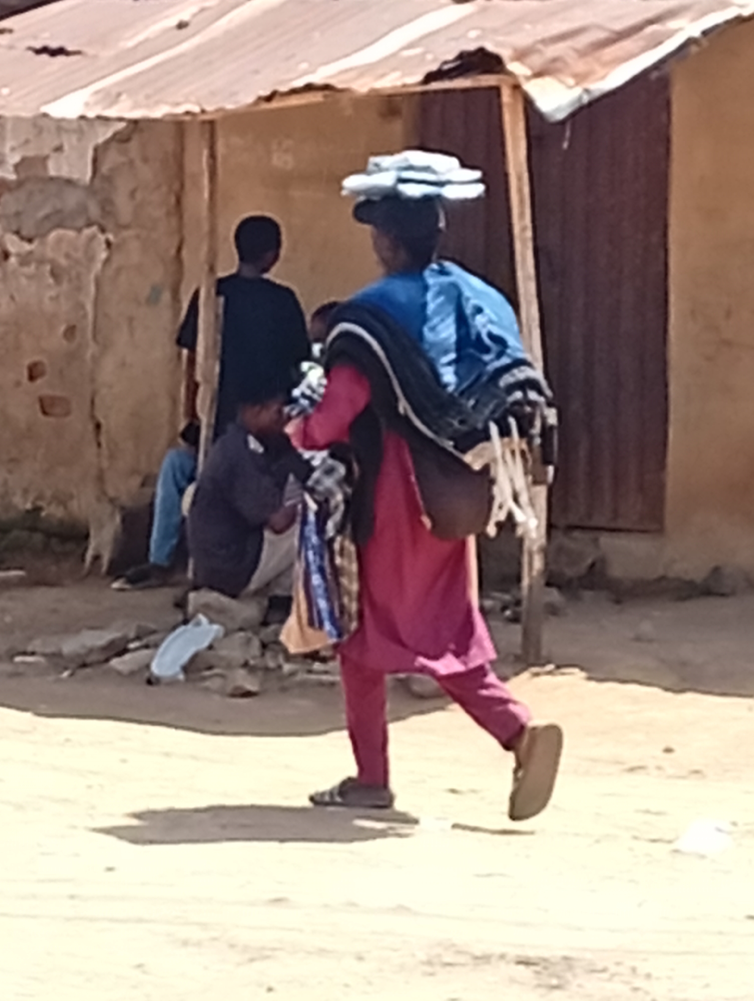 The image depicts an individual in Yelwa, Nigeria, walking on a dirt road in a residential area. The person is wearing a rose-colored tunic and trousers, sandals, carrying items on their head, a blue shawl around their shoulders, and a bag on their side. They appear to be carrying a child. Several other people are present in the background near a partially dilapidated building with a corrugated metal roof and wooden doors. Debris litters the ground. The environment is bright and sunny.