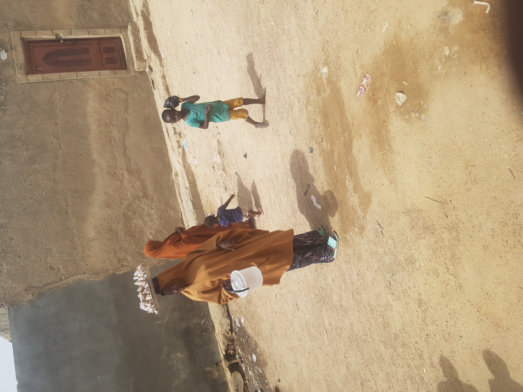 The image depicts an outdoor scene in Dutsinma B, Nigeria. Three people are visible: an adult woman dressed in a yellow robe with a white bucket, another adult in an orange robe, and a child wearing a green shirt and yellow shorts, carrying a black object. All the subjects are barefoot. The woman in yellow carries a tray covered in white objects, possibly food items, on her head. The ground appears to be dry and sandy with some scattered debris. There is a mud-brick structure with a window on the left side of the image.