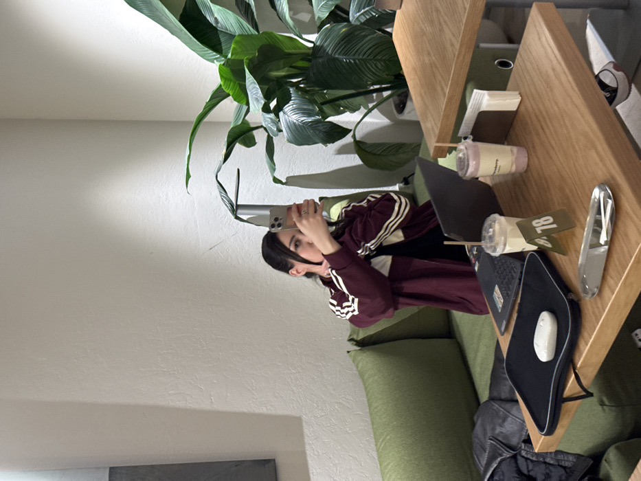 A young woman in a burgundy Adidas track jacket holds her smartphone and takes a selfie. She sits on a green couch next to a light wood desk. A large green plant stands nearby. A laptop, mouse pad, and drinks rest on the desk. There are also other objects like a clear paperweight, a notebook, and some cards. The room appears brightly lit with white walls.