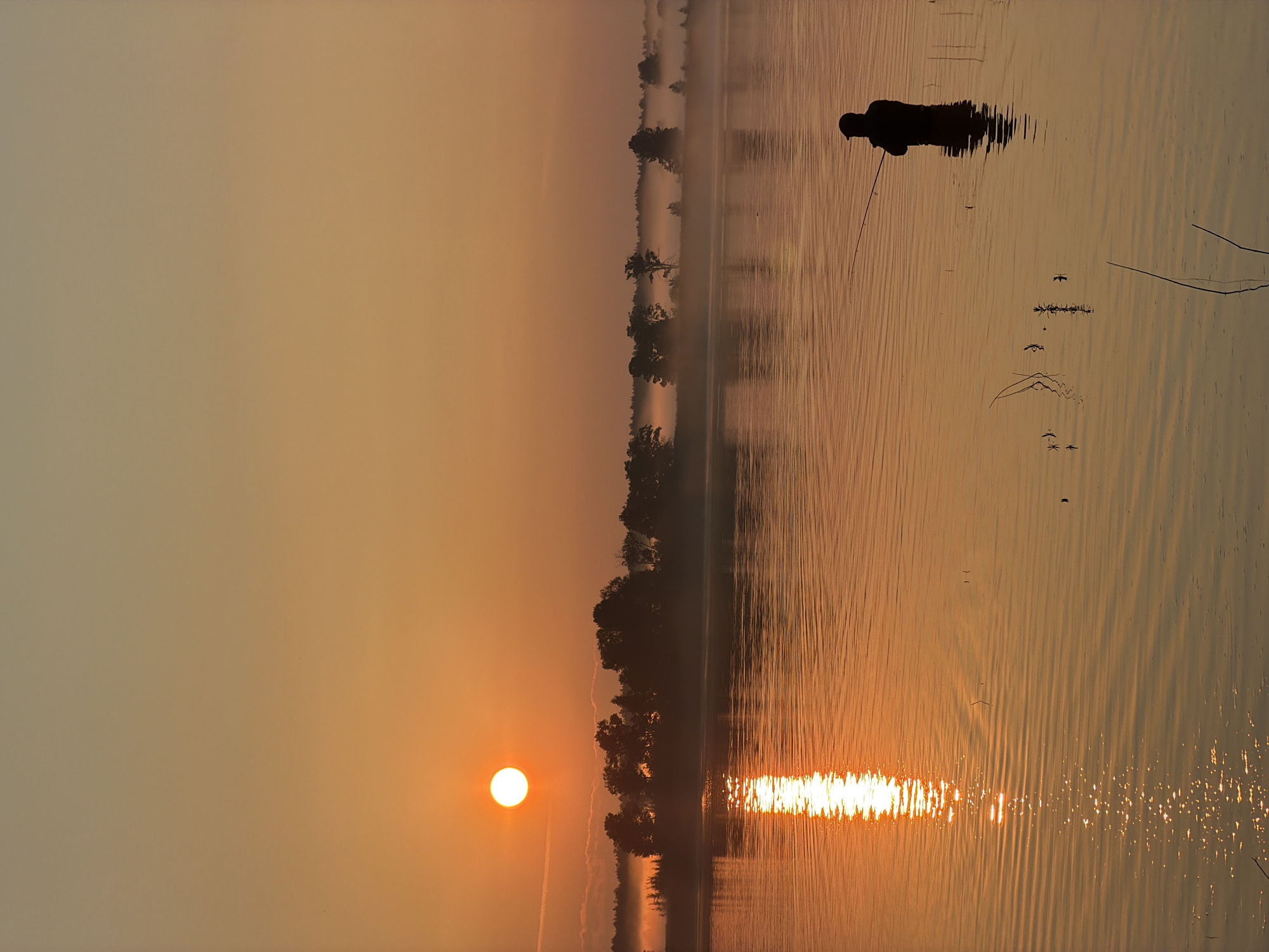 A solitary fisherman, silhouetted against the golden hues of a sunrise, stands in the shallow waters of a lake. The sun, a bright orb, casts a shimmering reflection across the rippling surface of the water. A distant shoreline is lined with trees and shrouded in mist, adding a sense of depth and tranquility to the scene. The sky transitions from a warm orange near the horizon to a pale peach at the top. The fisherman is positioned on the right side of the frame, facing away from the viewer, with a fishing rod in hand, actively engaged in casting a line. The overall atmosphere is serene and picturesque, evoking a sense of peace and quietude. The location is identified as the city of Chernushka, Russia.