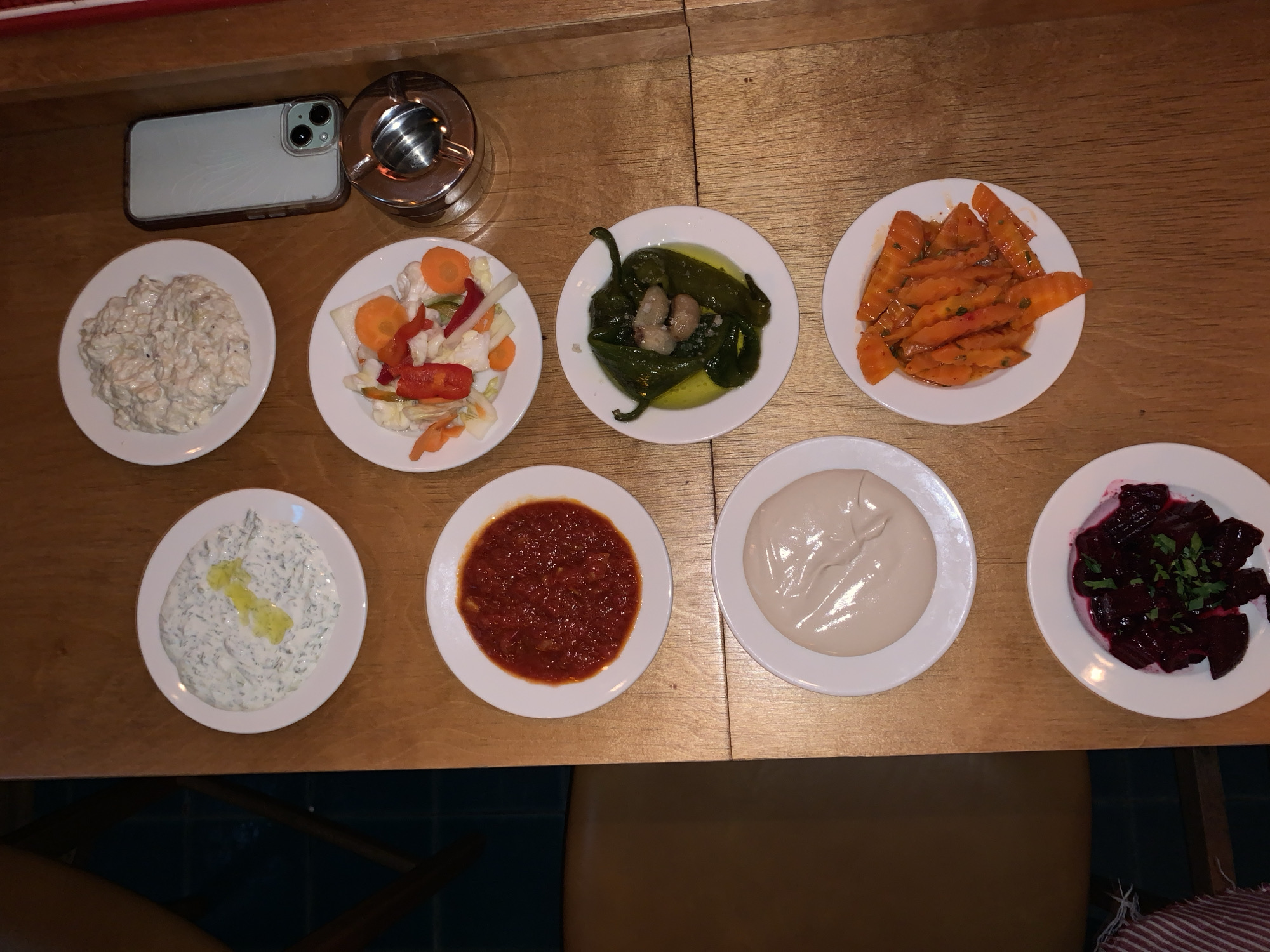 An overhead view captures a wooden dining table within a restaurant in Tel-Aviv, Israel. The table is occupied by nine small, round white plates, each containing a different food item, arranged in a grid-like pattern.
Specifically, the plates display:
* Top left: A creamy white dip with green flecks, garnished with a swirl of olive oil.
* Top right: A mound of creamy, off-white salad, possibly potato or chicken based.
* Middle left: A vibrant red, chunky relish or sauce.
* Middle right: Mixed pickled vegetables including orange carrot rounds, white cauliflower florets, and red pepper strips.
* Center right: Whole green peppers submerged in olive oil, topped with a few light-colored pieces of solid food and white crystalline flakes.
* Bottom left: A smooth, light beige-colored dip, likely tahini or hummus.
* Bottom center: Sliced orange vegetables, possibly carrots, in a reddish sauce.
* Bottom left (lowest): Diced dark red vegetables, likely beets, garnished with fresh green herbs.
To the upper right of the food arrangement, an iPhone with a clear protective case rests face down on the table, revealing its camera array. Directly below the phone is a cylindrical, metallic ashtray. The background shows a portion of a dark blue tiled floor and the leg of a wooden chair in the lower left, along with a glimpse of a red and white striped fabric. No individuals are visible in the image.
