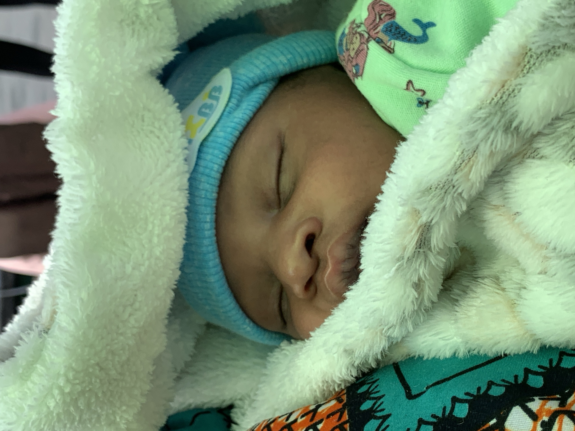 A baby is sleeping, wrapped in a white, fluffy blanket. The baby is wearing a light blue knit hat with a yellow and white applique. A green knit hat with mermaid designs is also visible. The background shows a multi-colored patterned fabric. The location is Ibasa, Nigeria.