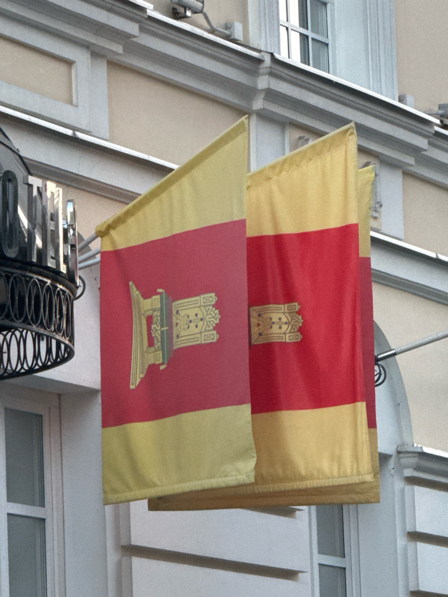 The image shows two flags hanging from a building in the city of Tver, Russia. The flags are rectangular and divided horizontally into two bands of color: red on top and yellow on the bottom. Each flag features a golden emblem in the center of the red band. The emblem appears to be a stylized representation of a crown or headdress, with intricate details and religious or heraldic symbols. The building behind the flags is light-colored, with visible architectural features such as windows and moldings. To the left of the flags, a dark, ornate metal structure is partially visible, which could be part of the building's facade or an entranceway. The flags are slightly furled, suggesting a gentle breeze. The overall scene depicts a patriotic or official display outside a building.