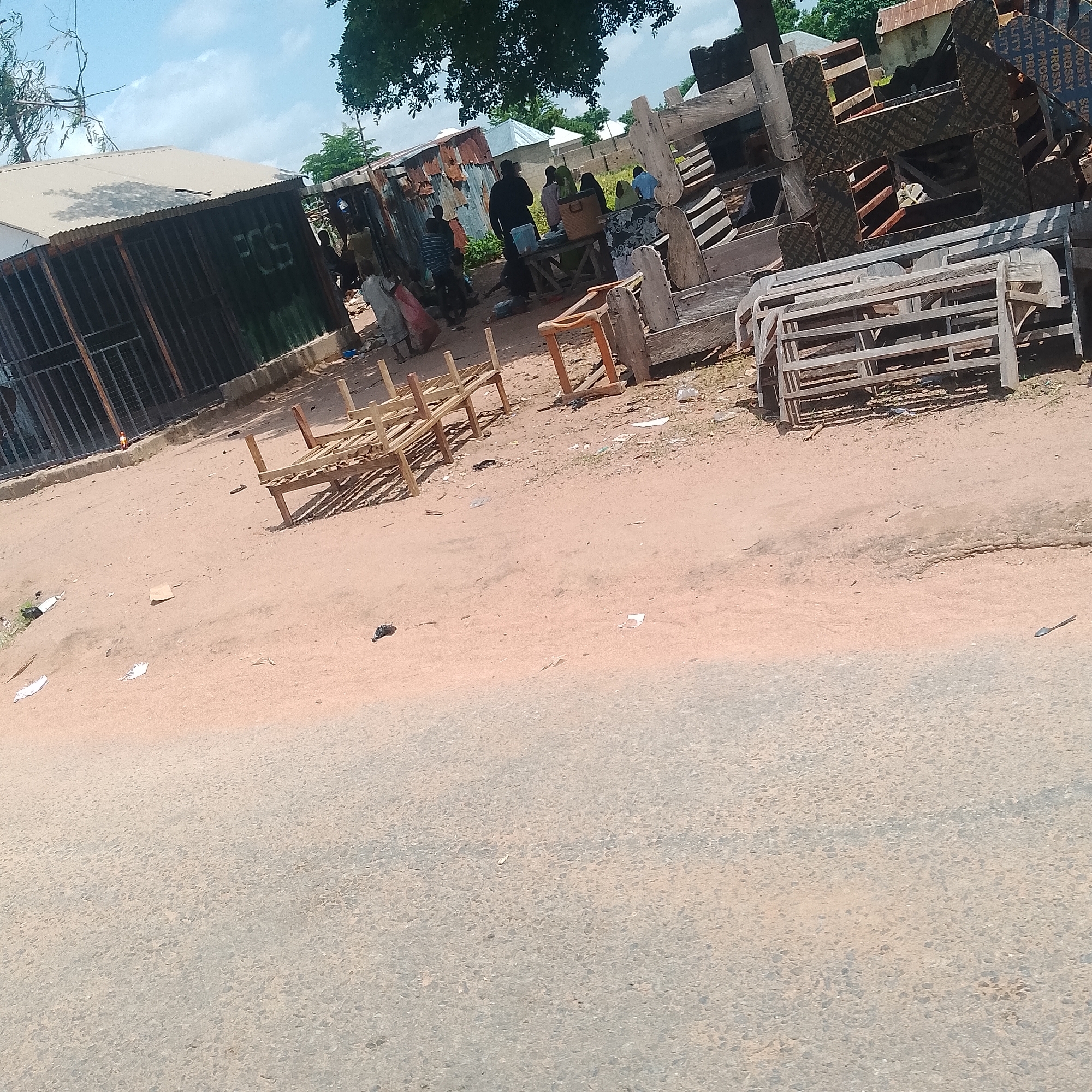 The image depicts an outdoor scene in Jalingo, Nigeria. In the foreground, a dusty, light-colored dirt road transitions to an asphalt surface. The background features a collection of market stalls and structures constructed from corrugated metal and wood. Several individuals are visible, some standing and others appearing to be interacting or engaged in activities within the market area. Scattered wooden furniture, possibly chairs and tables, are present in the scene, some appearing unfinished or stacked. The environment suggests a bustling, possibly informal, marketplace setting with a mix of permanent and temporary structures. The sky is partly cloudy.