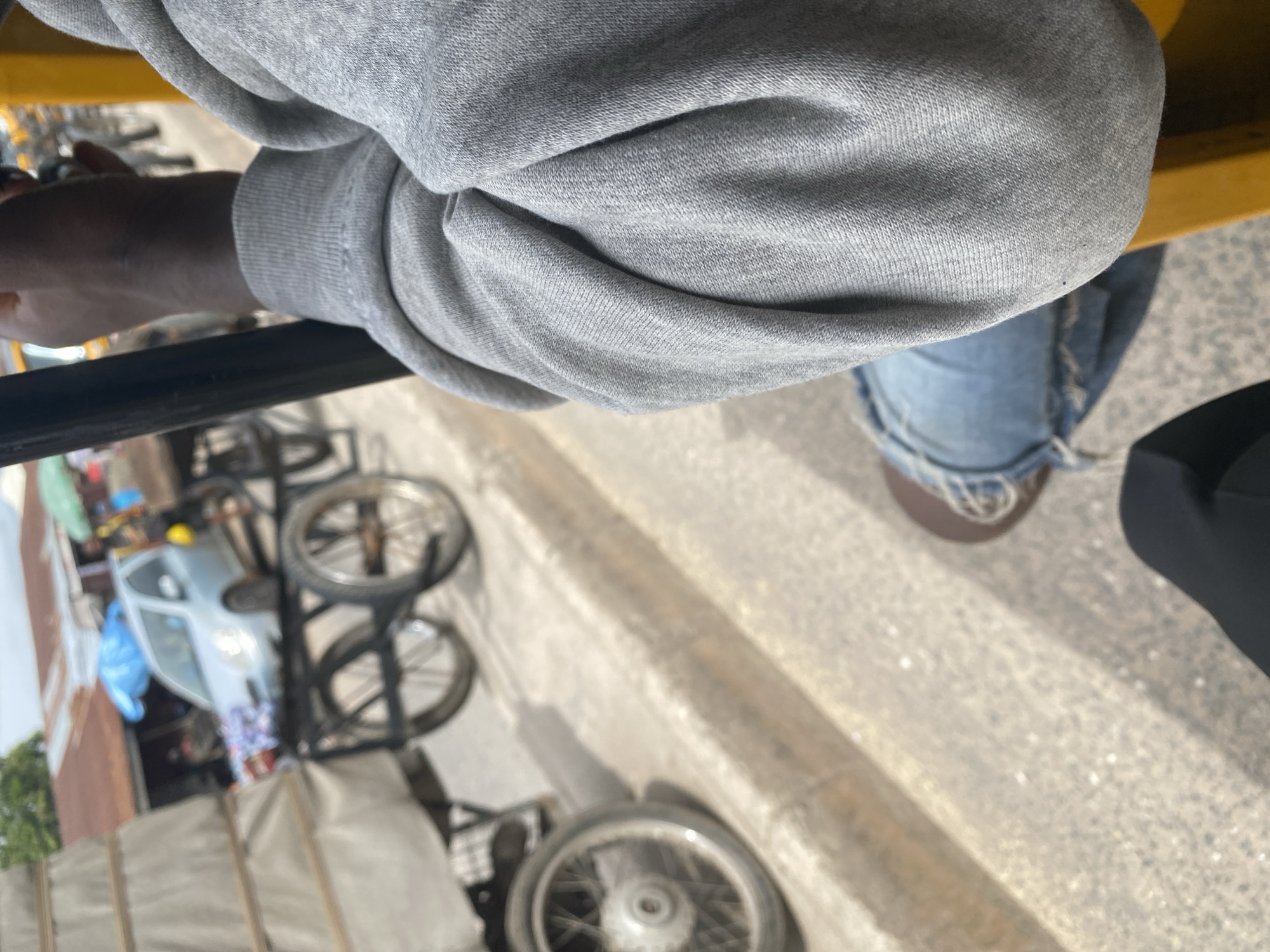 The image shows a close-up view of a person's arm and shoulder wearing a grey sweatshirt, resting on a black bar. The person is also wearing blue jeans and brown shoes. In the background, there are several bicycles, a light blue vehicle, and some market stalls. The scene appears to be outdoors, with a paved surface and a concrete curb. The lighting suggests it is daytime, possibly in a sunny environment. The overall impression is a street scene in a populated area, possibly a market or a busy street in Jalingo, Nigeria. The image is taken from a low angle, looking up towards the person's arm.