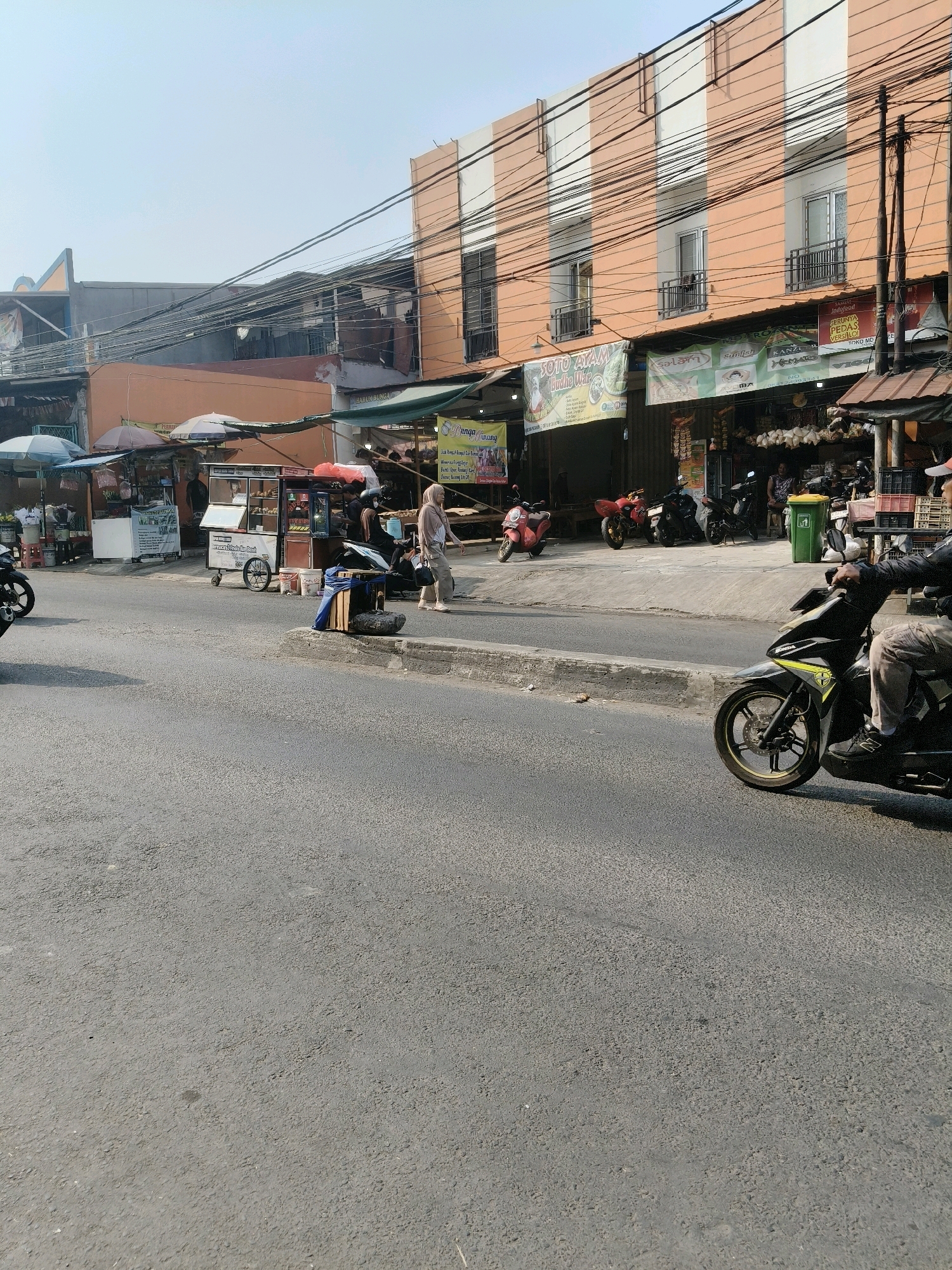 This is a medium-angle, eye-level, street-level, outdoor, daytime, medium shot of a sidewalk market in South Tangerang, Indonesia. The scene is dominated by a paved road in the foreground and a bustling street market in the background. The market features numerous stalls with umbrellas and awnings, selling various goods. Several motorcycles are parked and in motion throughout the scene.
In the foreground, a man in a black jacket and beige pants is riding a black and yellow motorcycle. He is facing to the right, with his hands on the handlebars. The road is paved and appears to be in moderate condition, with some cracks and patches visible.
The sidewalk market stretches across the background. There are several stalls displaying food and drinks, with umbrellas providing shade. One stall has a cart with a display case and a woman in a hijab standing nearby. Another stall has a banner that reads "SOTO AYAM Indohwar." Further down the street, there are more stalls and buildings with orange and white facades. A green trash can is visible near one of the stalls.
Several motorcycles are parked on the side of the road or in front of the stalls. There are also people walking and interacting at the market. The sky is clear and bright, indicating a sunny day. The overall atmosphere is that of a lively urban market scene.