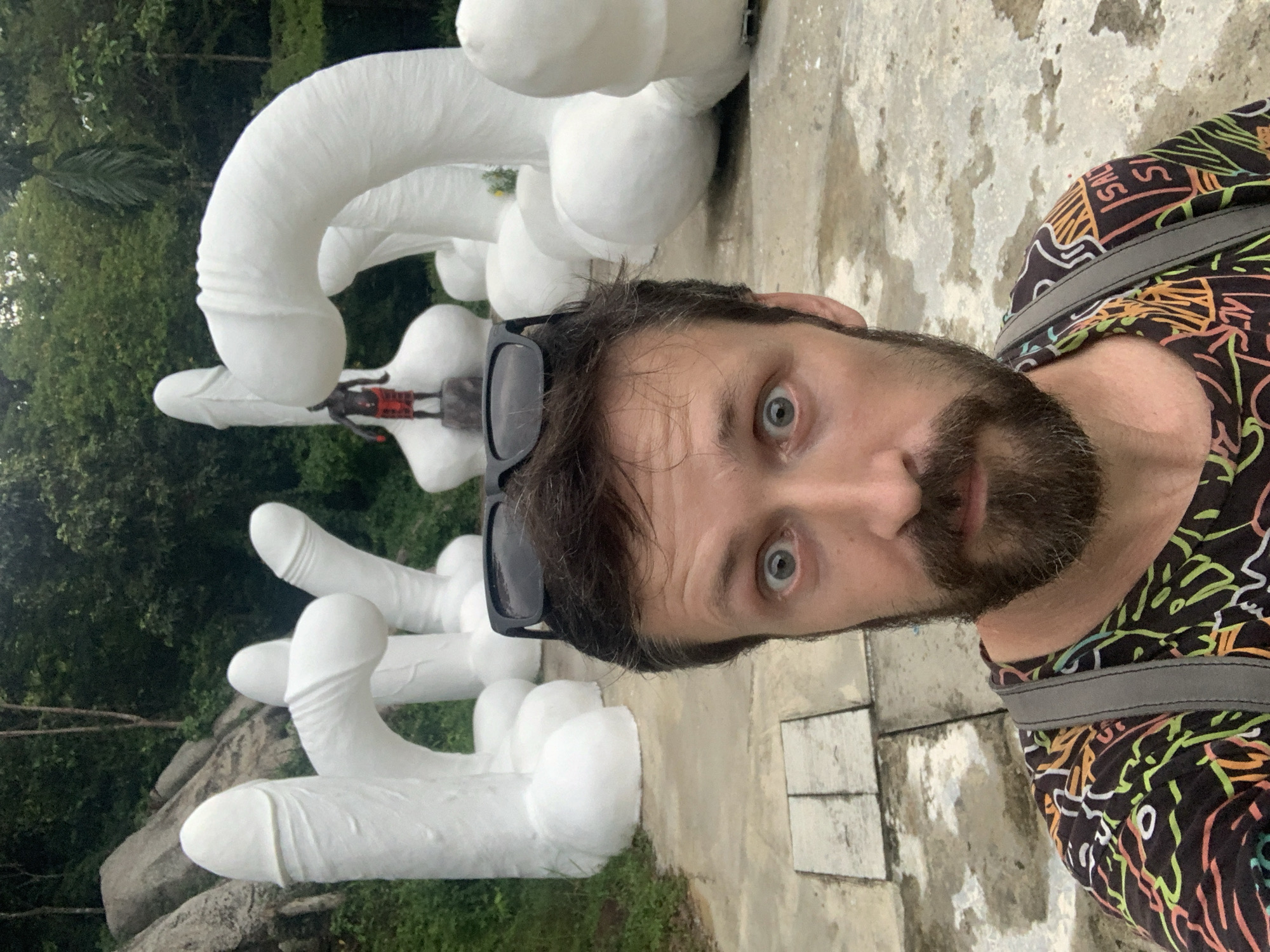 A man takes a selfie, captured from a slightly high and rotated angle. He has a beard and dark hair, with sunglasses resting on his head. His blue eyes are wide open, conveying an expression of surprise or amusement, and he looks directly at the camera. He wears a dark shirt with vibrant, abstract patterns in green, orange, white, and yellow, and a gray backpack strap is visible on his shoulder.
In the background, behind the man, are numerous large, white, phallic-shaped sculptures. These objects vary in size and curve, resembling oversized penises. One small, dark-skinned figure, possibly a doll or statue, dressed in red shorts and a dark top, is seated on one of these sculptures. The setting is outdoors, with lush green trees and foliage visible in the distance behind the sculptures. The ground is a textured, light-colored surface, possibly concrete or paved. The scene is brightly lit, suggesting it is daytime. This location, given the context of Baan Hua Thanon, Thailand, is likely a traditional fertility shrine. No discernible text is visible in the image.