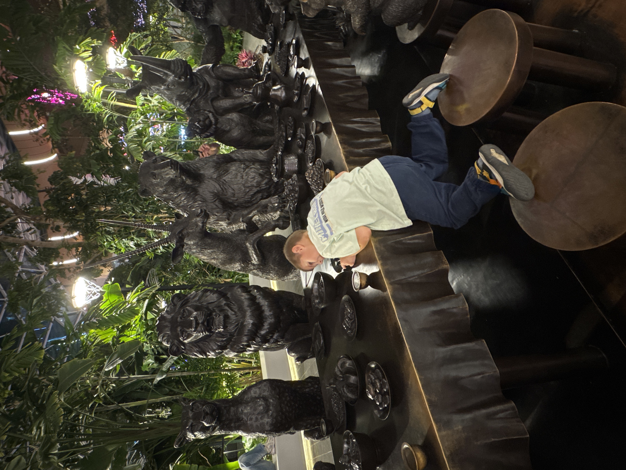 In Doha, Qatar, a young boy wearing a light green t-shirt and dark blue pants is leaning over a dark, rustic table laden with numerous small dark bowls and plates, some appearing to contain food items. He is actively engaged with something on the table. The table is set in a dimly lit, indoor garden environment. In the background, several large, dark, animal sculptures are visible, including what appears to be a lion, a bear, a rhinoceros, and other mammals. Lush green foliage and overhead lighting create an atmospheric backdrop, suggesting this is a curated exhibition or display space, possibly within a botanical garden or a themed area of a public attraction in Doha. The boy's body language and focus suggest he is intently examining the items on the table.