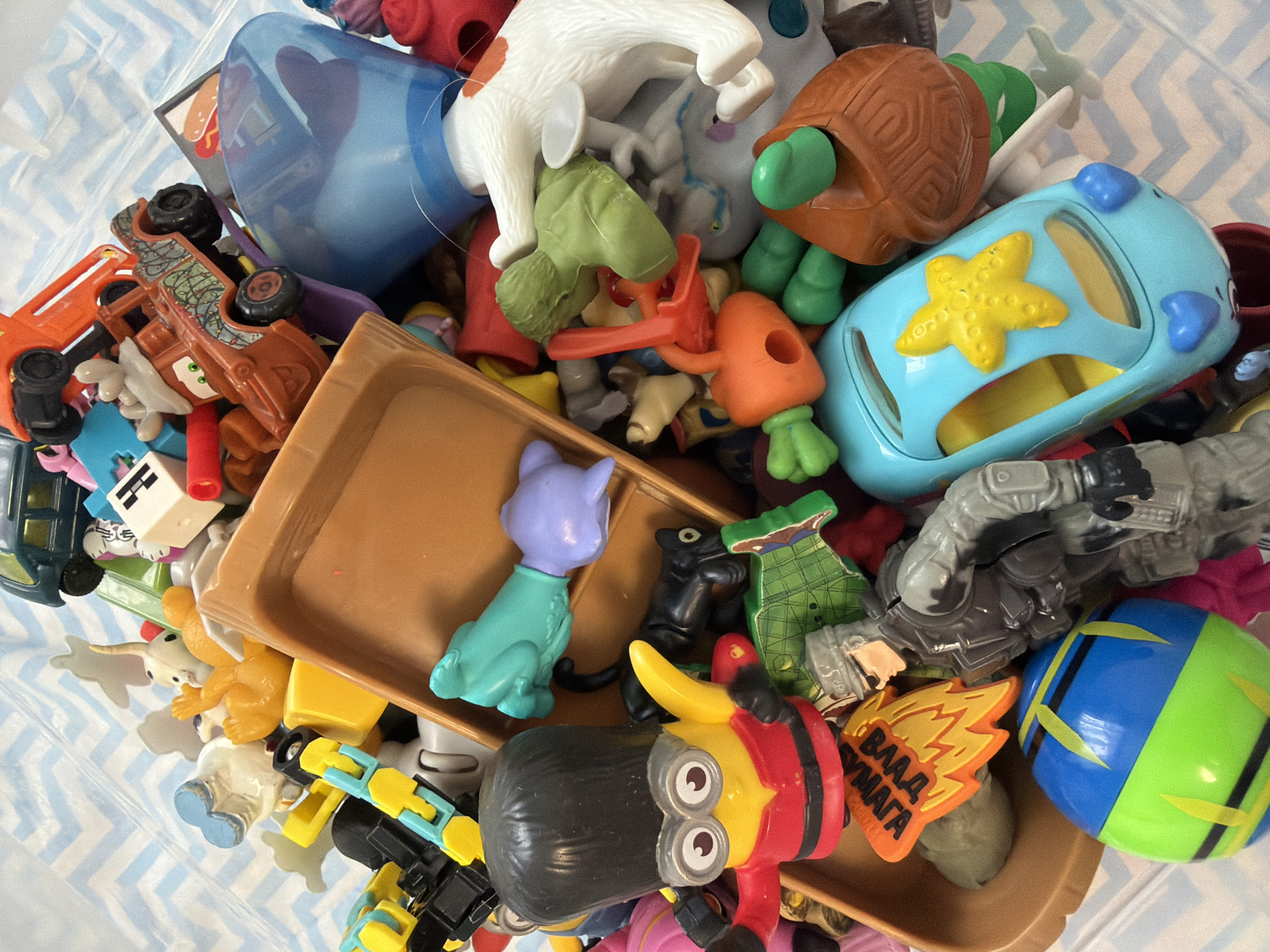 A high-angle shot shows a jumble of colorful plastic toys filling a tan plastic tray and overflowing onto a light-blue-and-white patterned surface. The toys include a blue toy car with a yellow starfish, a brown turtle toy, a Minion figure, a green-and-blue ball, a light-blue cat figure, a gray action figure, and assorted other small plastic toys. The location is not identifiable as being in Votkinsk, Russia. There are no people visible.