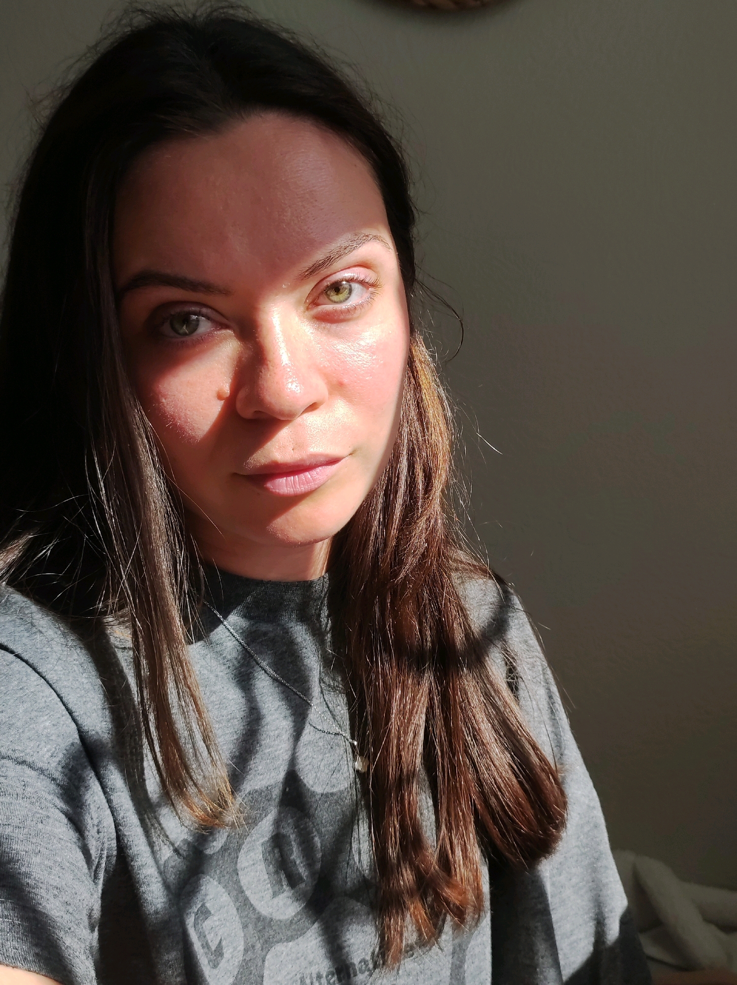 A woman with long, dark brown hair and green eyes is captured in a close-up, head-and-shoulders shot. She is wearing a grey t-shirt with a subtle, light grey graphic design. The lighting is dramatic, with strong sunlight casting sharp shadows across her face and the background, which appears to be an interior wall. The sunlight highlights her forehead, cheekbones, and the bridge of her nose, creating a strong contrast with the shadowed areas. Her expression is neutral, with her gaze directed towards the camera. The image does not provide any specific visual cues to determine the location within Chaikovsky, Russia.