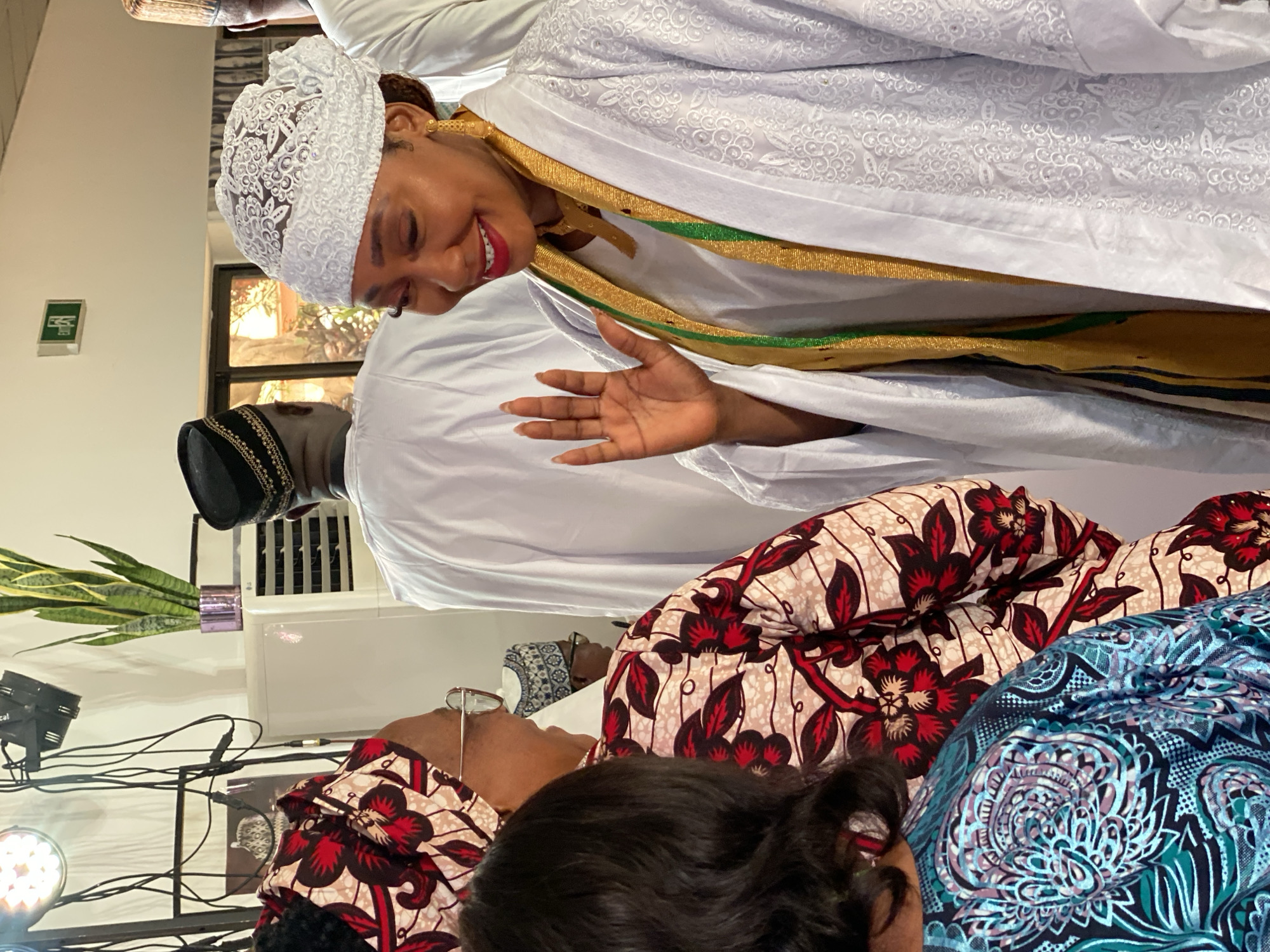 The image appears to be taken indoors, possibly in a gathering space, in Kwarau, Nigeria. A woman in the top portion of the image is wearing a white lace head covering and a white robe with gold and green trim. She is smiling and has her hand raised in a waving gesture. Behind her, partially obscured, is a man in a black hat and white robe. Below them, there are two women sitting and/or lying down. One woman is wearing a floral patterned top, with a red floral pattern over light skin and the second woman is wearing a dark green floral patterned top over darker skin. The background shows various lights and wires, a potted plant and an air conditioner.
