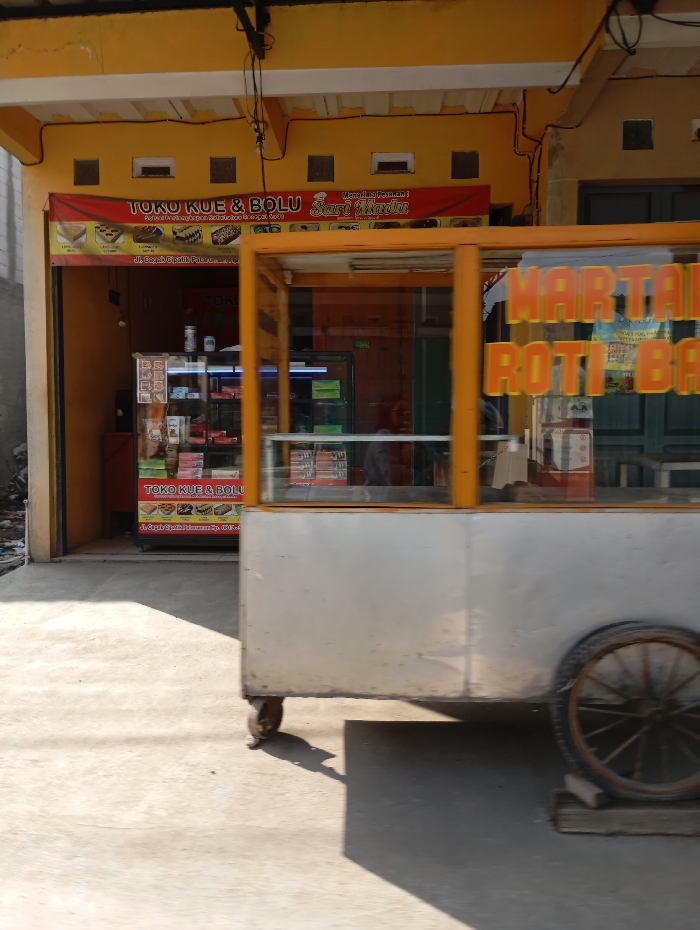 The image depicts an outdoor commercial scene in Cipatik, Indonesia, specifically at a location identified as Jl. Cigajah Cipatik Pasiraman Km. 0,5. The primary subject is a single-story building with a bright yellow facade and white trim, featuring an open storefront. A prominent red banner above the entrance identifies the establishment as "Toko Kue & Bolu Sari Mardiana," displaying illustrations of various baked goods.
Inside the shop, multiple glass display cases filled with packaged food items and a refrigerator are visible. In the immediate foreground, a light-colored, glass-paneled street food cart partially obscures the shop's interior. Orange lettering on the cart's front glass, partially obscured by glare, indicates "MARTABAK" and "ROTI BAKAR." The cart rests on small front caster wheels and a larger, spoked wooden wheel at its rear, which is elevated by wooden blocks. The ground in the foreground is a flat concrete surface. Strong shadows are cast from the cart and the building, indicative of direct sunlight. Black electrical conduits are observable on the building's exterior facade. No individuals are present within the frame.