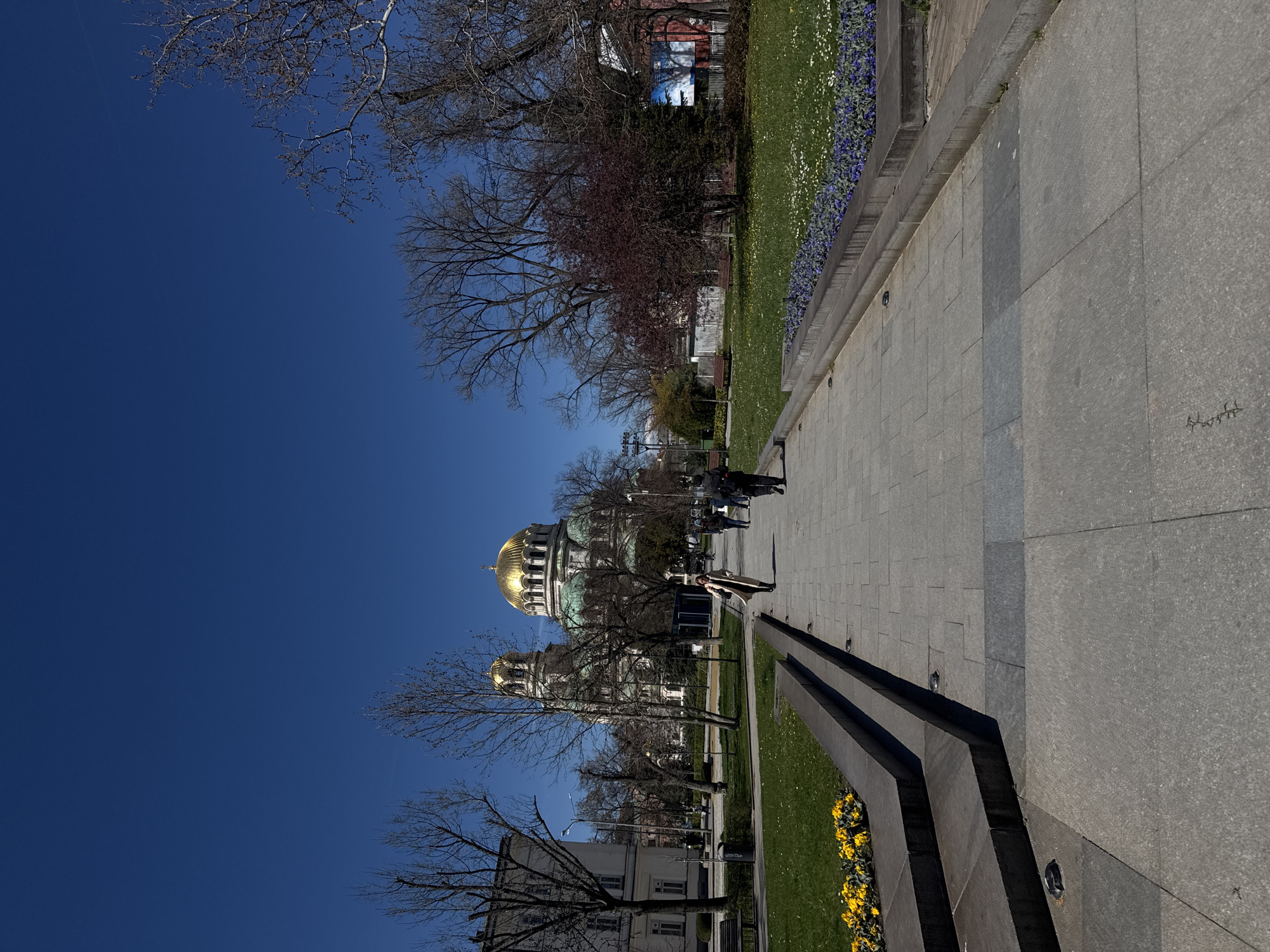 The image shows a sideways view of St. Alexander Nevsky Cathedral in Sofia, Bulgaria, under a clear blue sky. The cathedral's golden domes are prominent. In the foreground is a paved walkway alongside a grassy area with flower beds. Several people are walking along the pathway. Trees with bare branches are interspersed throughout the scene. Buildings can be seen in the background. The environment appears to be a well-maintained public space in an urban setting.