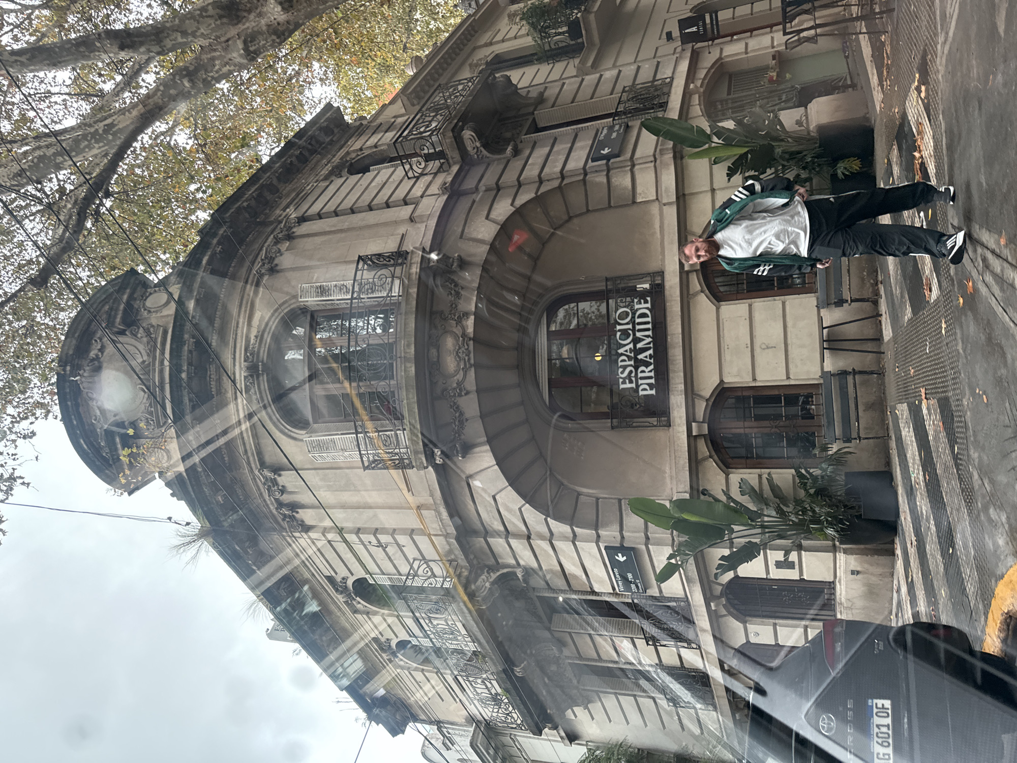 The photo depicts a street scene in Buenos Aires, Argentina. The foreground shows a reflection in a car window, revealing a partial view of the dashboard and the license plate "AG 601 OF." A man stands on the sidewalk, wearing a white t-shirt, a green and white Adidas jacket, and black pants with white stripes. He is facing the camera with his head tilted. Behind him is a corner building with ornate architectural details, including arched windows, iron balconies, and decorative moldings. The building's facade is gray. A sign with the words "ESPACIO PIRAMIDE" (Pyramid Space) is visible above a doorway. Plants in large containers flank the building's entrance. A directional sign points towards a location listed as "TANTI 2200-2299." A tree with bare branches is visible in the upper left corner of the frame.