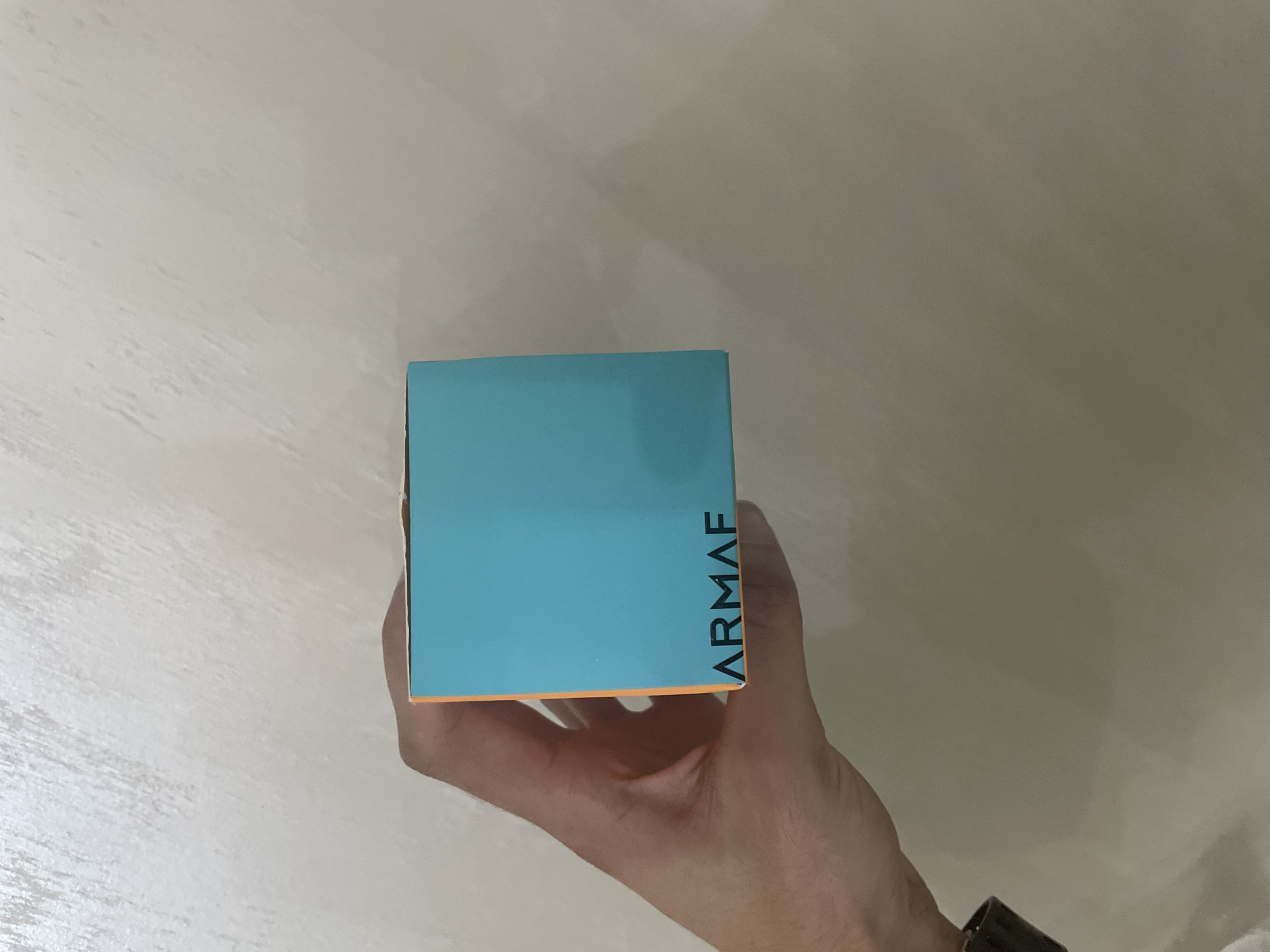 A left hand holds a teal and orange rectangular box against a textured, light-toned wall. The visible face of the box is solid teal with the brand name 'ARMAAF' printed in black capital letters on the bottom left. An orange side panel is partially visible on the left. The hand, with a light to medium skin tone, grips the box from the left side and beneath, revealing a dark wrist strap near the bottom. The background wall is uniformly lit, displaying an off-white or light beige color with subtle, irregular surface texture.