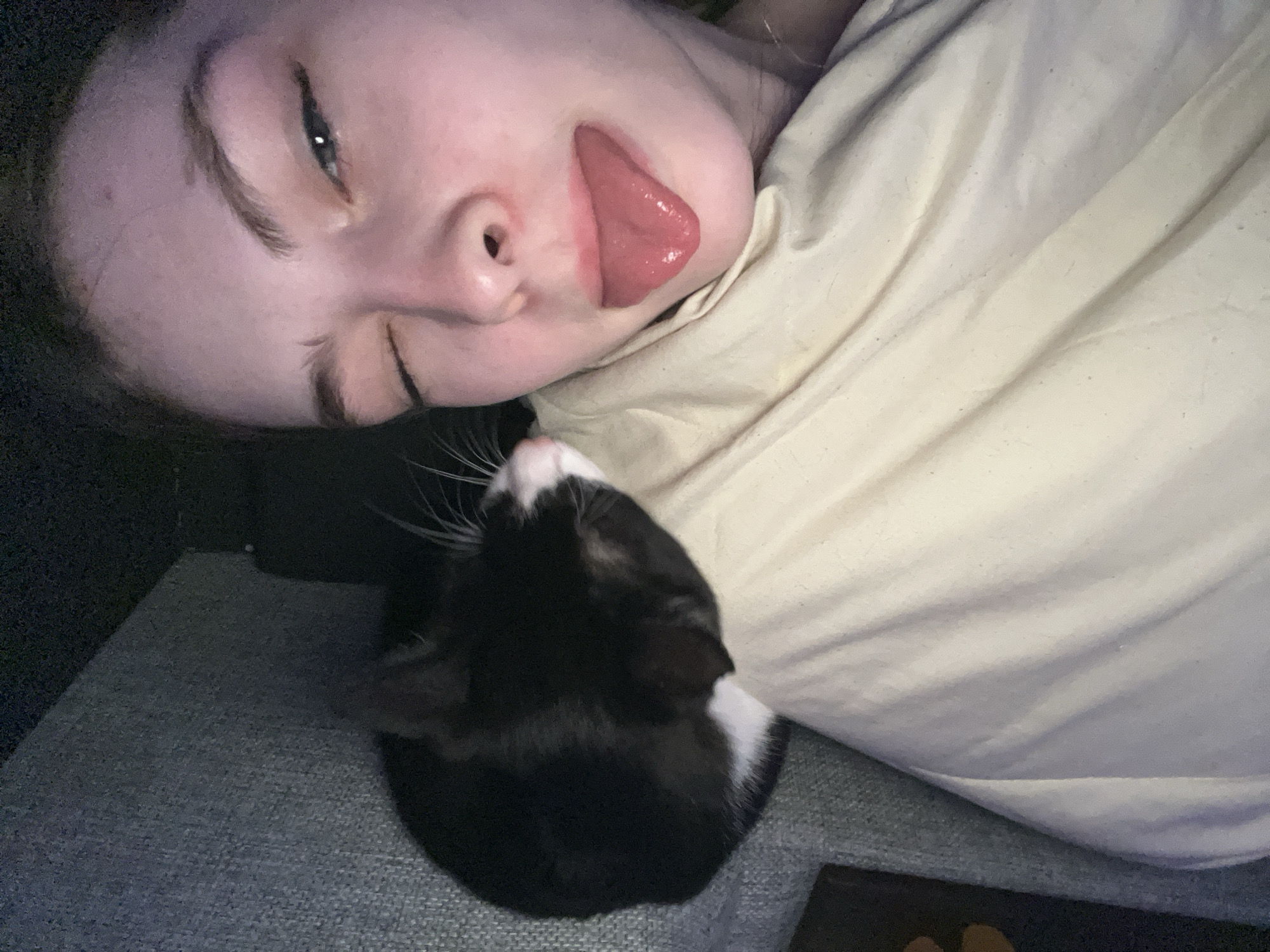 A person is lying on their side, sticking their tongue out, next to a black and white cat. They are lying on a gray textured surface next to a rumpled white fabric. It is not possible to identify the location as Saint Petersburg, Russia based on the image.