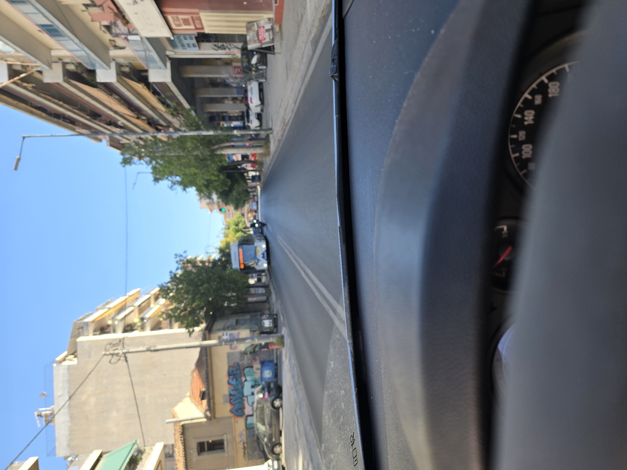 The image captures a view from the interior of a car looking out through the windshield onto a street in Athens, Greece. The street is lined with multi-story buildings, some with balconies and awnings, and trees. A white bus is visible in the distance, driving down the street. Cars are parked along the side of the road, and there is some graffiti on a building on the left. The sky is clear and blue. The car's dashboard is partially visible in the foreground, including a speedometer. The overall scene suggests a daytime urban environment.