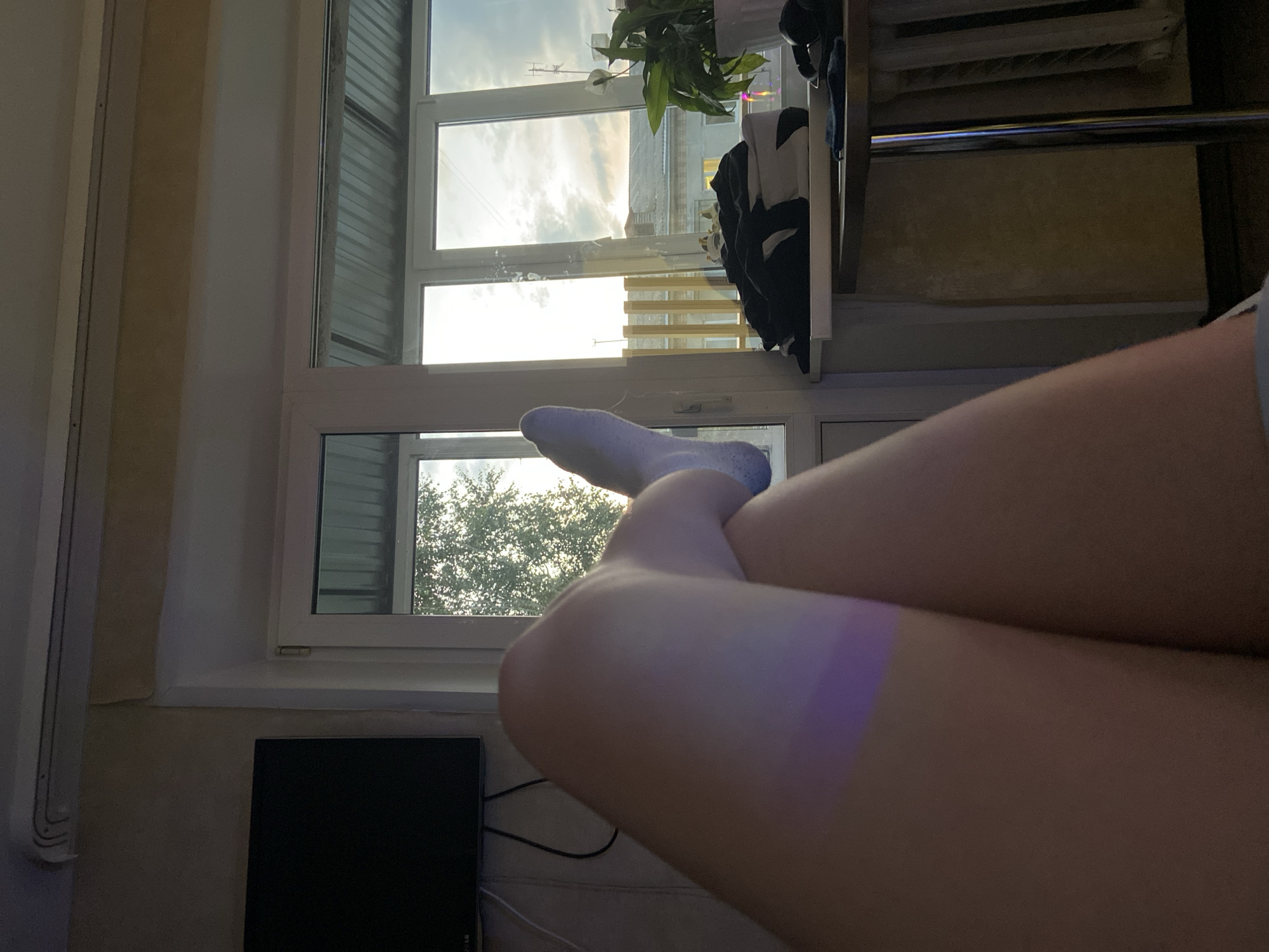 The image shows a person's legs in the foreground, crossed, with one foot wearing a light blue sock. A purple light is cast on the left leg. In the background, a window with multiple panes reveals an outdoor scene with trees and buildings under a cloudy sky. A television is mounted on the wall to the left. A shelf on the right holds several folded clothes and a potted plant. The overall setting appears to be an indoor living space with a view of an urban environment. The city of Beloretsk, Russia, is mentioned in the prompt but there are no visual elements in the image that confirm this specific location.
