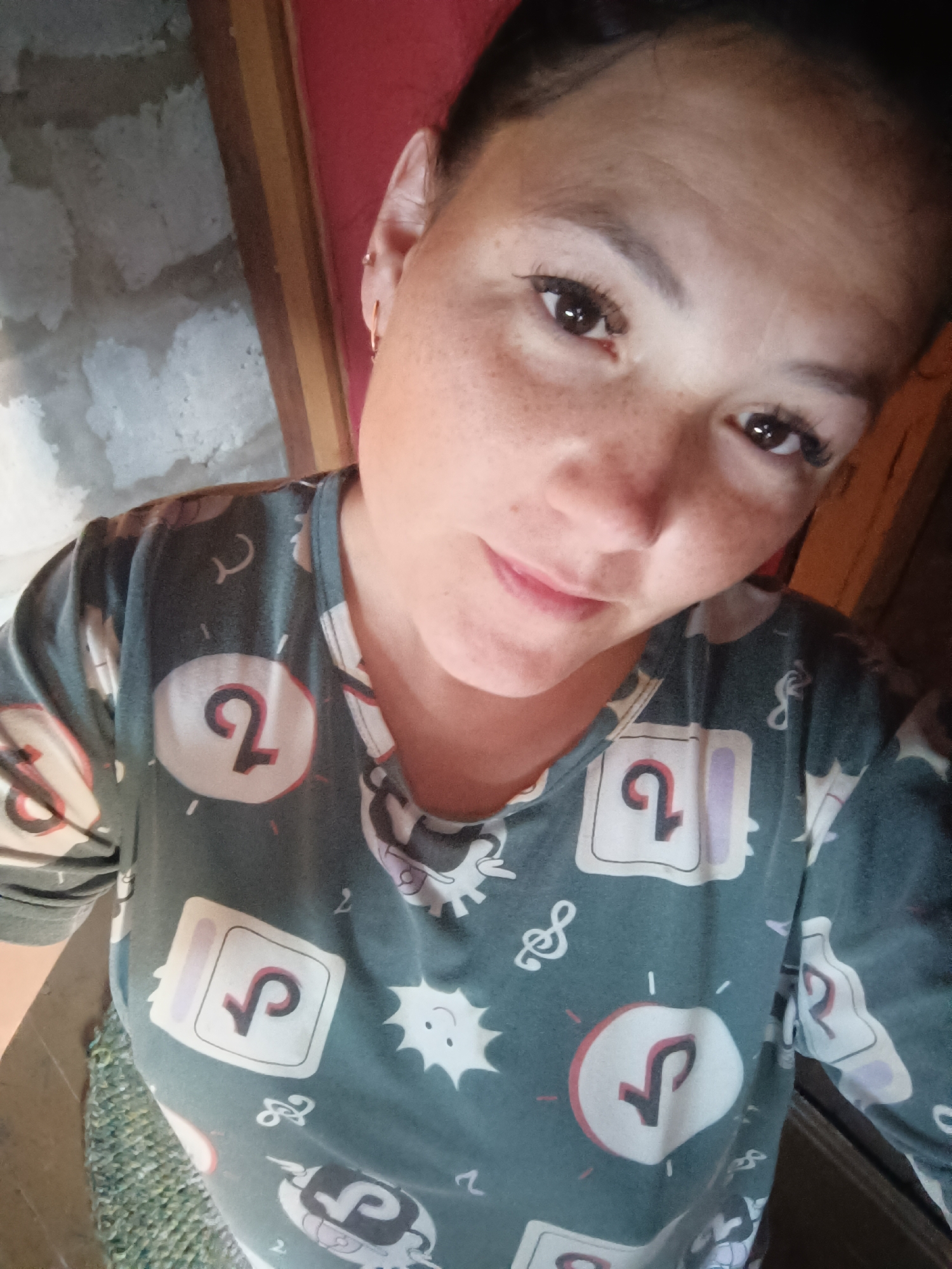The image is a close-up, selfie-style photograph of a woman with fair skin and brown eyes, looking directly at the camera. She has freckles across her nose and cheeks. She is wearing a dark teal-colored t-shirt with a repeating pattern of white squares containing a stylized "2" or a TikTok logo, as well as other small graphics like musical notes and cartoon suns. Her hair is dark brown and pulled back. She is wearing small hoop earrings in her left ear. The background is indistinct and appears to be an indoor setting, with hints of a concrete or brick wall on the left and a wooden structure or door on the right. There is no discernible information about the city of Karsha, Russia in the image.