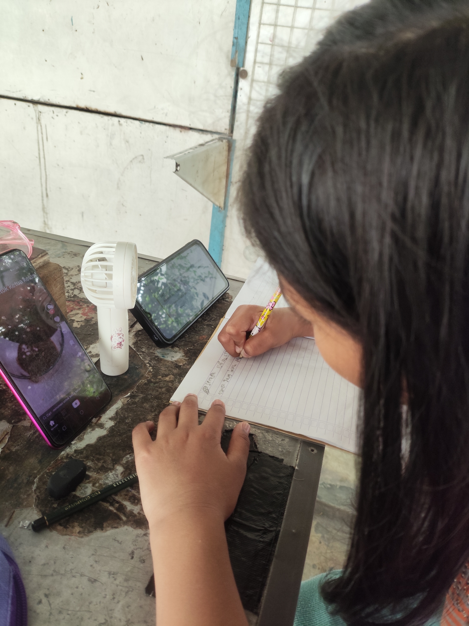The image depicts a close-up of a young person's hands as they write in a notebook. The person has long, dark hair that is visible from behind, obscuring their face. They are holding a pencil in their right hand and are actively writing. To the left of the notebook, there are two smartphones, one with a pink case and the other black, positioned on a worn, textured surface that appears to be a table or desk. A small white portable fan stands between the phones. An eraser and a black pen with text are also present on the surface. The background consists of a white, possibly metal or painted wall with some marks and a blue vertical element. The overall scene suggests a study or work session. This image was taken in Pekanbaru, Indonesia.