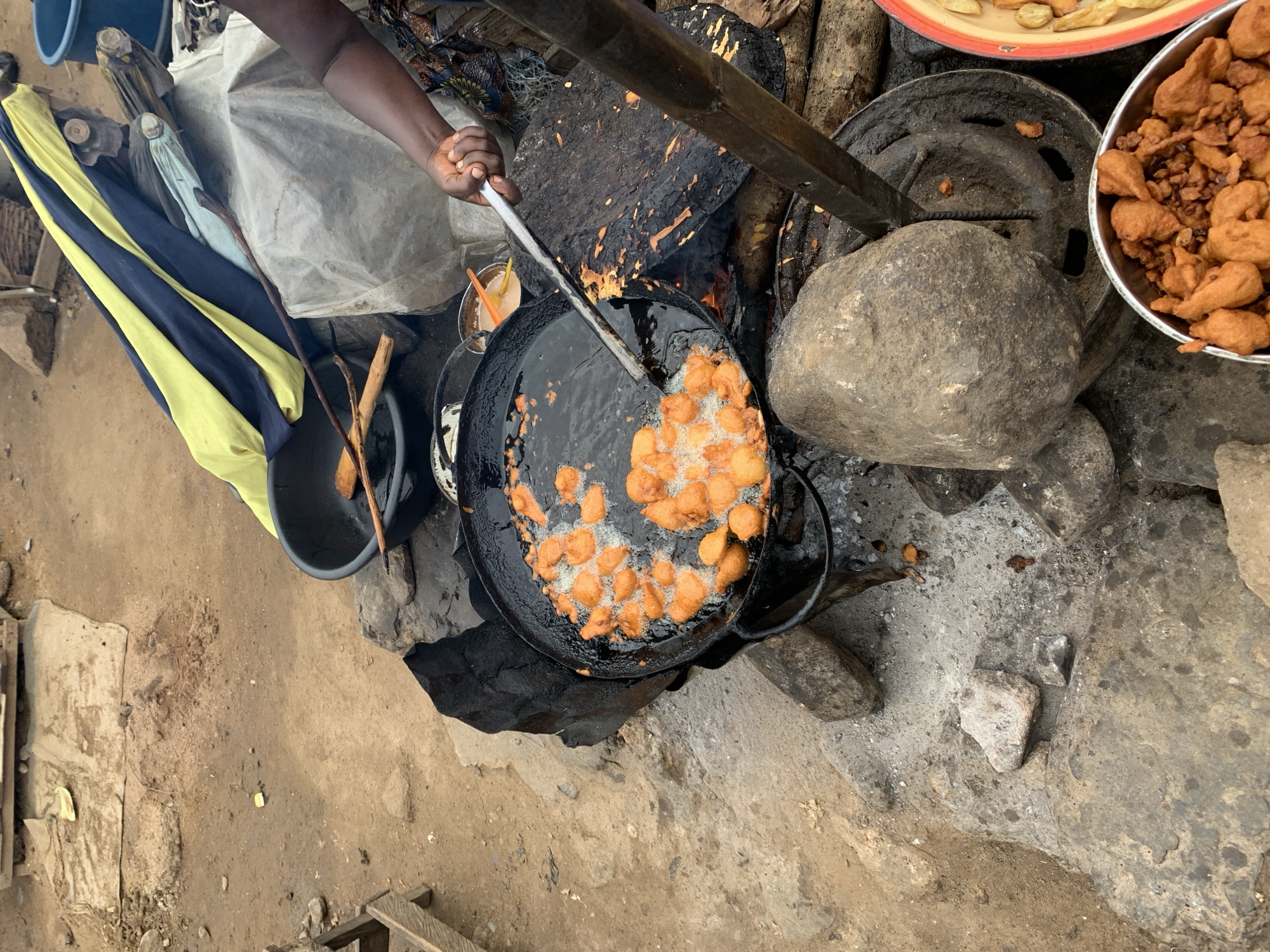 The image depicts an outdoor cooking scene in Rafin Par, Nigeria. A dark-skinned individual's right arm and hand are visible on the right side of the frame, holding a long, silver-colored metal utensil over a large, black, circular frying pan. The pan is filled with bubbling oil and numerous small, irregularly shaped, golden-orange fried items, actively cooking. The pan is supported by a makeshift cooking setup, including large, rough-hewn rocks and what appears to be a metal stand. An open fire, fueled by wood, is visible beneath and to the right of the pan, with visible flames and glowing embers. A large, dark, burnt wooden plank or log rests beside the fire.
To the bottom right, a silver metal bowl contains a pile of various fried food items, including golden fritters and smaller, darker, crispier pieces. Adjacent to it, a yellow and orange plastic plate or bowl holds more fried items, resembling chips and fritters. Further back, near the person's hand, a small, light brown liquid or batter is visible in a white bowl with an orange plastic spoon.
The ground is composed of light brown dirt or sand, interspersed with grey concrete or hardened soil patches and scattered small rocks and debris. In the upper left background, a simple wooden structure is partially visible. The upper right background features a dark blue plastic basin containing wooden utensils, and behind it, a folded yellow and dark blue striped fabric, along with other lighter-colored fabrics and a grey tarp covering an obscured object. A dark blue plastic bucket is also visible in the far upper right. The scene is illuminated by natural daylight.