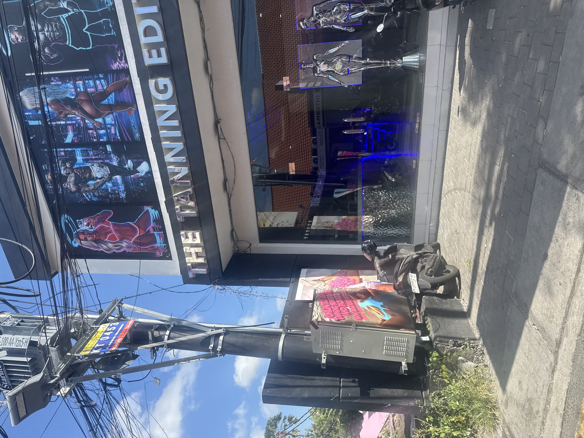 This media file captures a bright, sunny street scene in North Kuta, Indonesia, likely during midday or early afternoon due to the strong shadows cast on the paved sidewalk. The main subject is a building housing "THE TANNING EDIT," which features large window displays showcasing stylized, muscular figures, including both male and female forms with a cyberpunk aesthetic. The shop windows below these displays feature chrome, mannequin-like figures resembling futuristic robots.
In front of the building, a dark-colored scooter with the license plate "DK 4095 FAE" is parked on the sidewalk, next to a stand-up poster advertising "NO MINIMUM TANNING." A prominent utility pole dominates the left side of the frame, laden with a dense tangle of power lines and cables. Attached to this pole are a street light, partially obscuring text like "Starlite" and "-100-AA-Yzn5-H," and a banner displaying "K10453" alongside a red and blue design. An electrical box is also mounted on the pole. The sky is clear and blue with scattered white clouds. Green foliage grows at the base of the utility pole and scooter. Reflected in the shop's windows, the word "Global" can be seen in reverse.