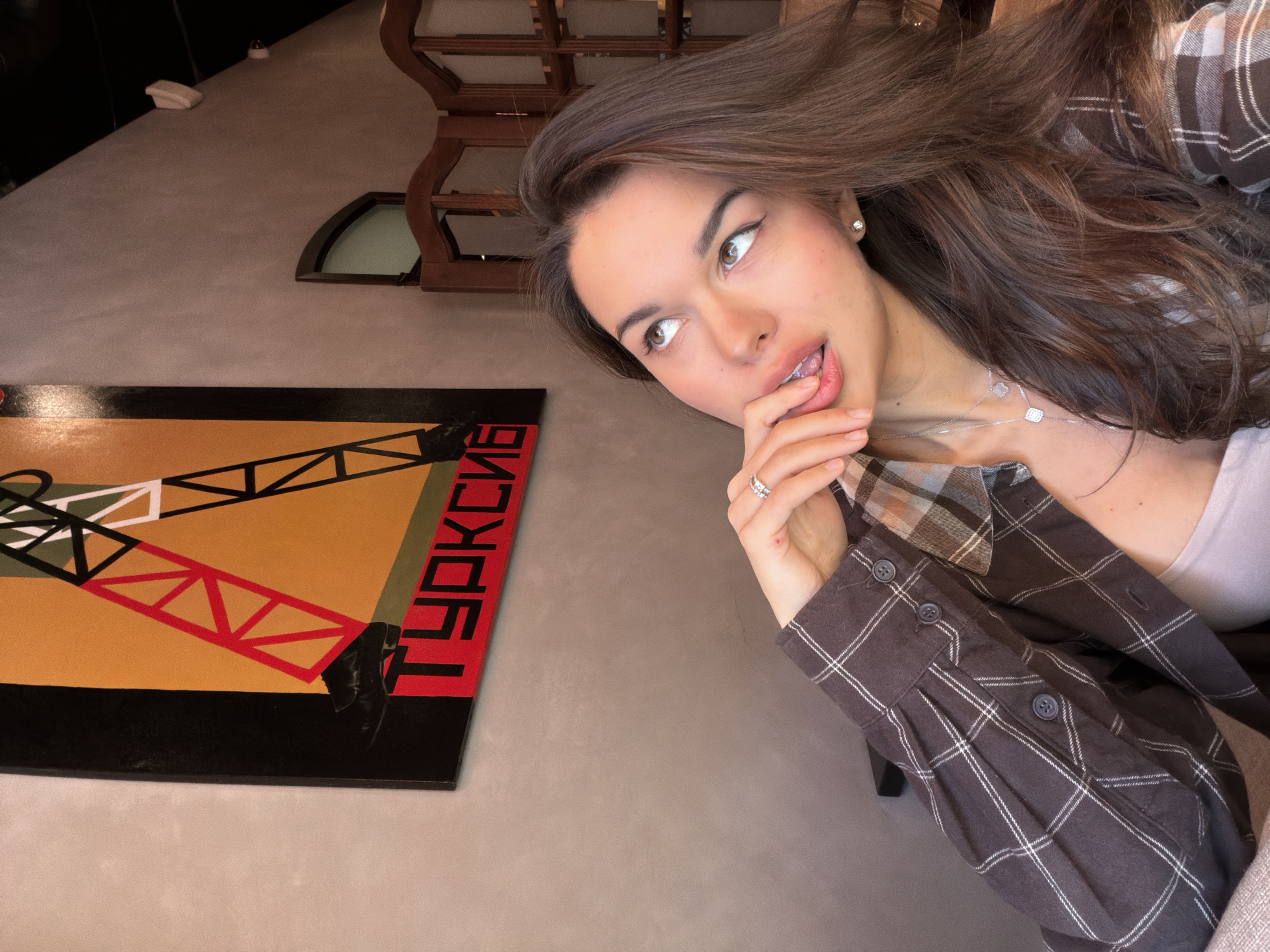 A woman is photographed indoors. Her long brown hair cascades over her shoulders and she wears a plaid shirt. Her left hand is raised to her face with her finger near her mouth, and she gazes off to the side. She wears a necklace and earrings. Next to her is an abstract painting with geometric shapes in red, black, yellow, and green. A word in red lettering is visible on the painting. Based on the text on the painting and other visual cues, the location is in Krasnoyarsk, Russia. A partial view of a wooden structure, possibly a chair or piece of furniture, is in the background.