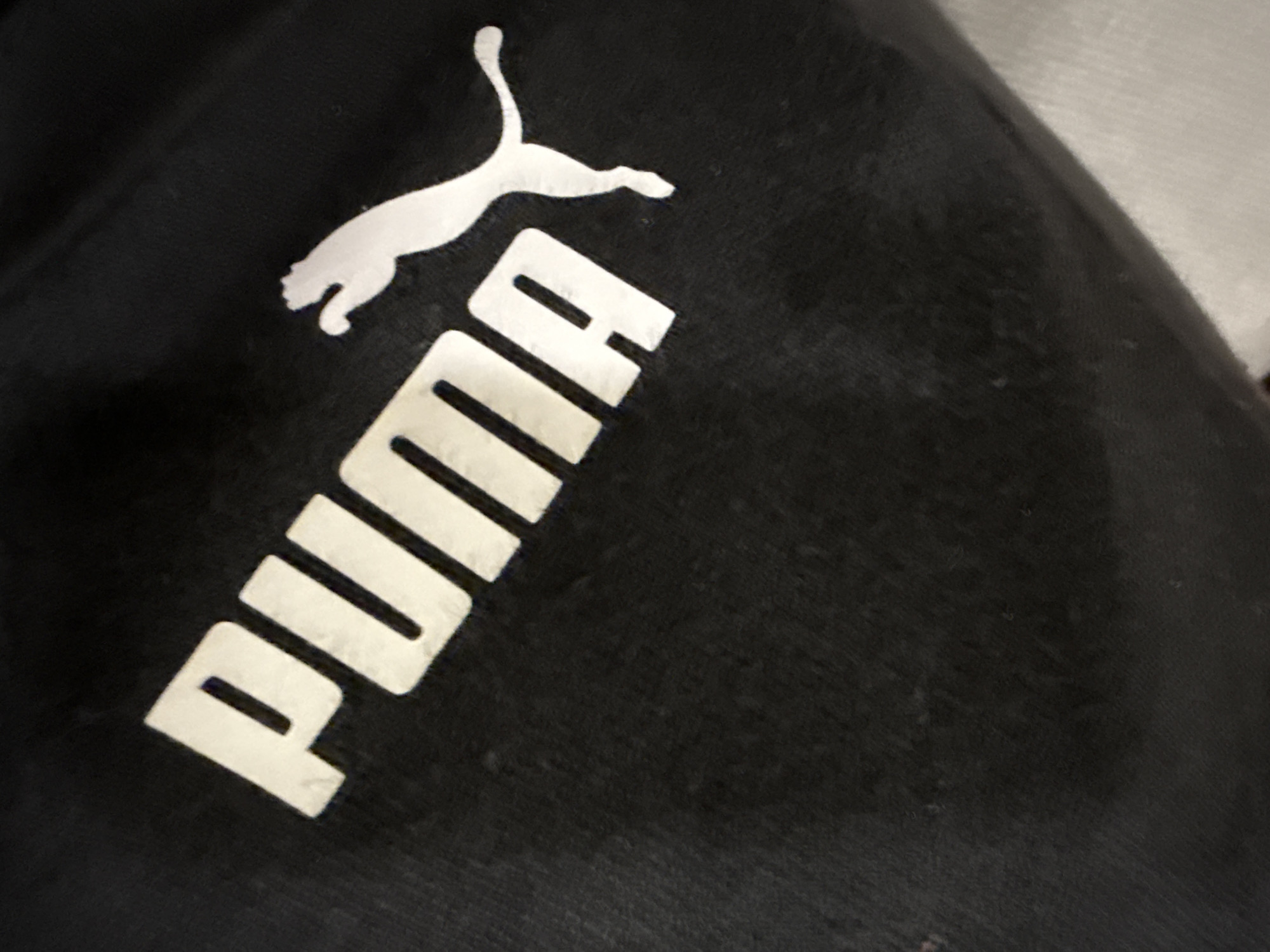 The image shows a black Puma-branded garment, likely a piece of clothing such as sweatpants or a sweatshirt. The white Puma logo, consisting of the leaping cat and the brand name "PUMA" in block letters, is prominently displayed on the garment. The fabric appears slightly worn and textured. There are no people visible in the frame. The location is not identifiable from the image.