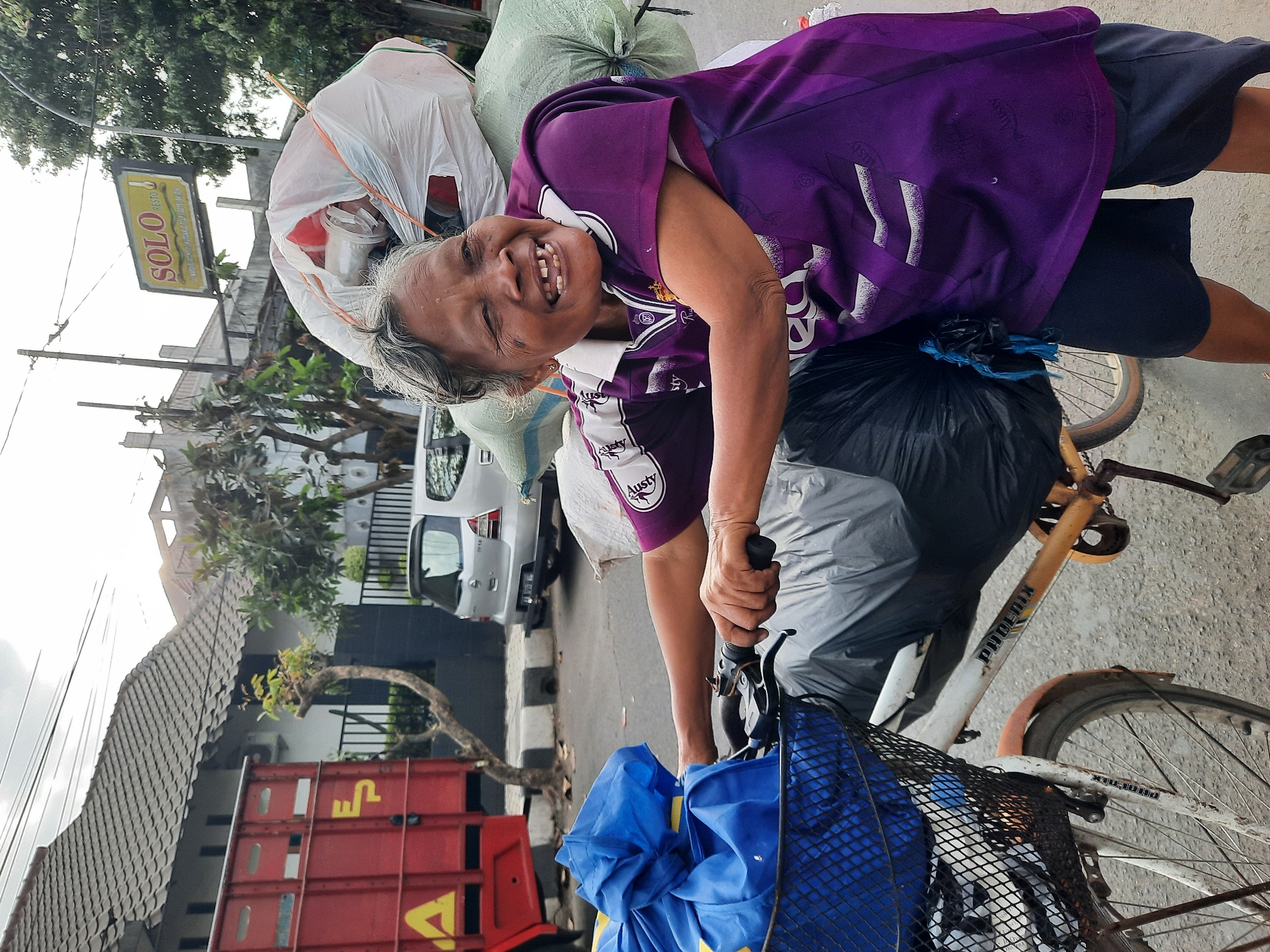 An elderly woman with graying hair and weathered skin is smiling broadly while riding a bicycle laden with large bags of what appears to be recycling. She is wearing a purple t-shirt with white accents and dark shorts. The bicycle has a basket in front filled with a blue bag. In the background, a red truck, a silver car, and buildings are visible. A sign for "SOLO RESTO" is also present. The scene suggests a street in the city of Kutoarjo, Indonesia. The woman's expression and the context of her load suggest she is a waste collector or scavenger. The overall atmosphere is one of daily life in an urban environment.