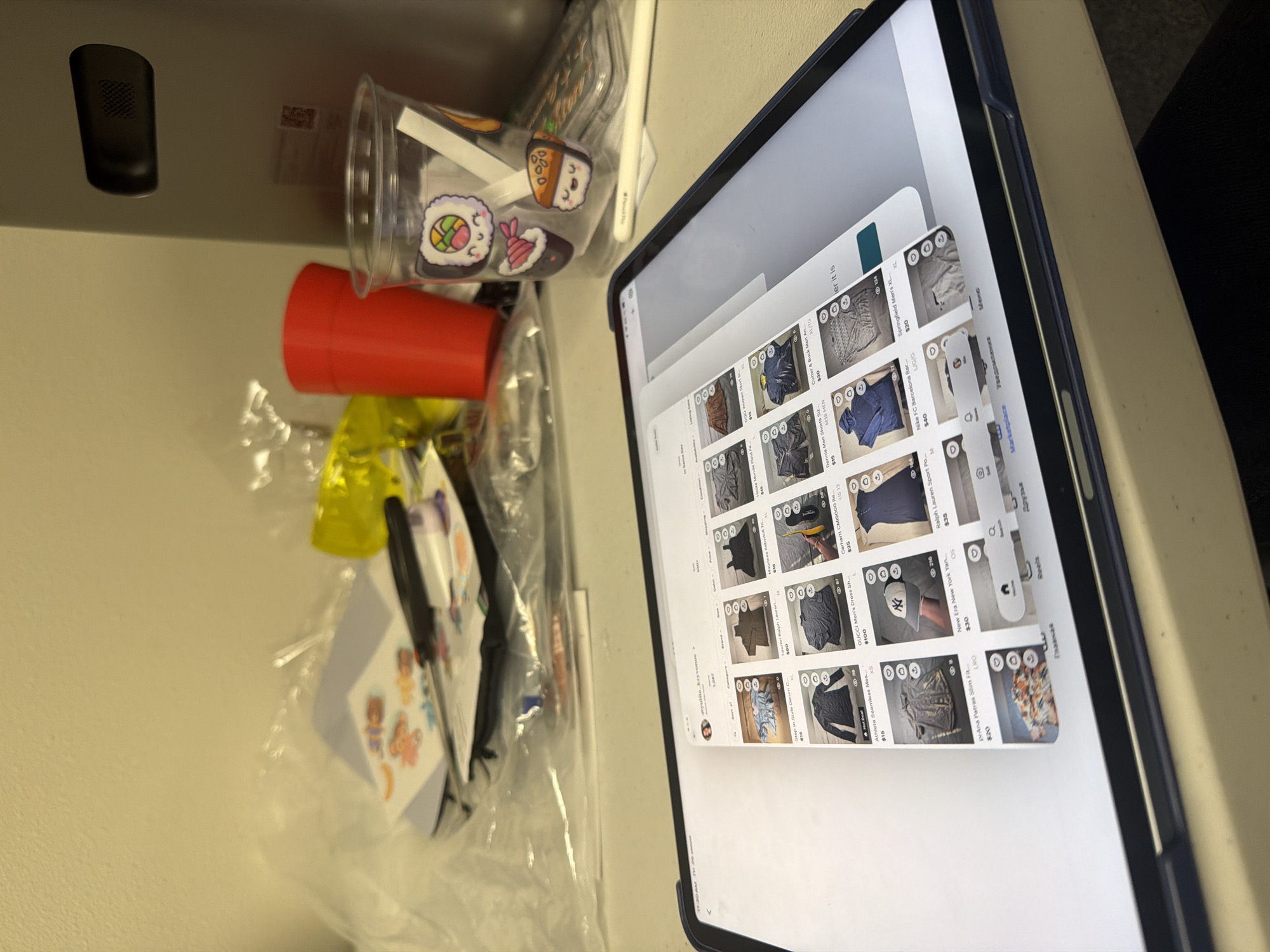 This media file depicts a still life composition featuring everyday objects. In the foreground, a tablet device displays a grid of clothing items for sale, suggesting an online shopping or marketplace application. The screen shows various apparel, including shirts, jackets, and a baseball cap, with prices ranging from $10 to $100. The tablet is placed on a light-colored surface, possibly a desk or table.
To the left of the tablet, several items are scattered. A clear plastic cup filled with small stickers, some resembling sushi, is visible. Next to it, a bright red cylindrical object, possibly a cup or container, lies on its side. Below these items, a yellow plastic bag and more translucent plastic packaging are present, hinting at a storage or organization context. A black electronic device, possibly a speaker or sensor, is mounted on a dark vertical surface in the upper left quadrant.
The lighting suggests an indoor setting, likely during the daytime, given the ambient light illuminating the scene. There are no people visible in the frame, and the overall mood is neutral and utilitarian. The visible text on the tablet screen is predominantly product descriptions and prices, with some interface elements like "Marketplace" and "Главная" (Russian for "Main"). The setting appears to be a room in Beaverton, United States of America.