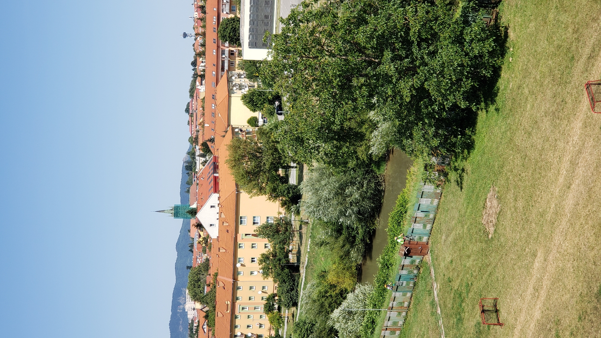 The image captures a view of the city of Prešov, Slovakia, likely from an elevated position looking towards a residential area and a river. In the foreground, a grassy field contains a small, red soccer goal. Adjacent to the field is a wooden fence bordering a small river or canal, with two individuals visible near the fence. One person is standing, and another is on a bicycle. Lush green trees and vegetation line the riverbank.
Beyond the river and trees, a street is visible with apartment buildings and houses. The apartment buildings are multi-story structures with orange tiled roofs and light-colored facades, featuring numerous windows and satellite dishes. In the background, a church spire, partially obscured by scaffolding, rises above the rooftops. Further in the distance, rolling hills are visible under a clear blue sky. To the right, a water tower and other city structures can be seen. The overall scene depicts a sunny day in a suburban or semi-urban environment.