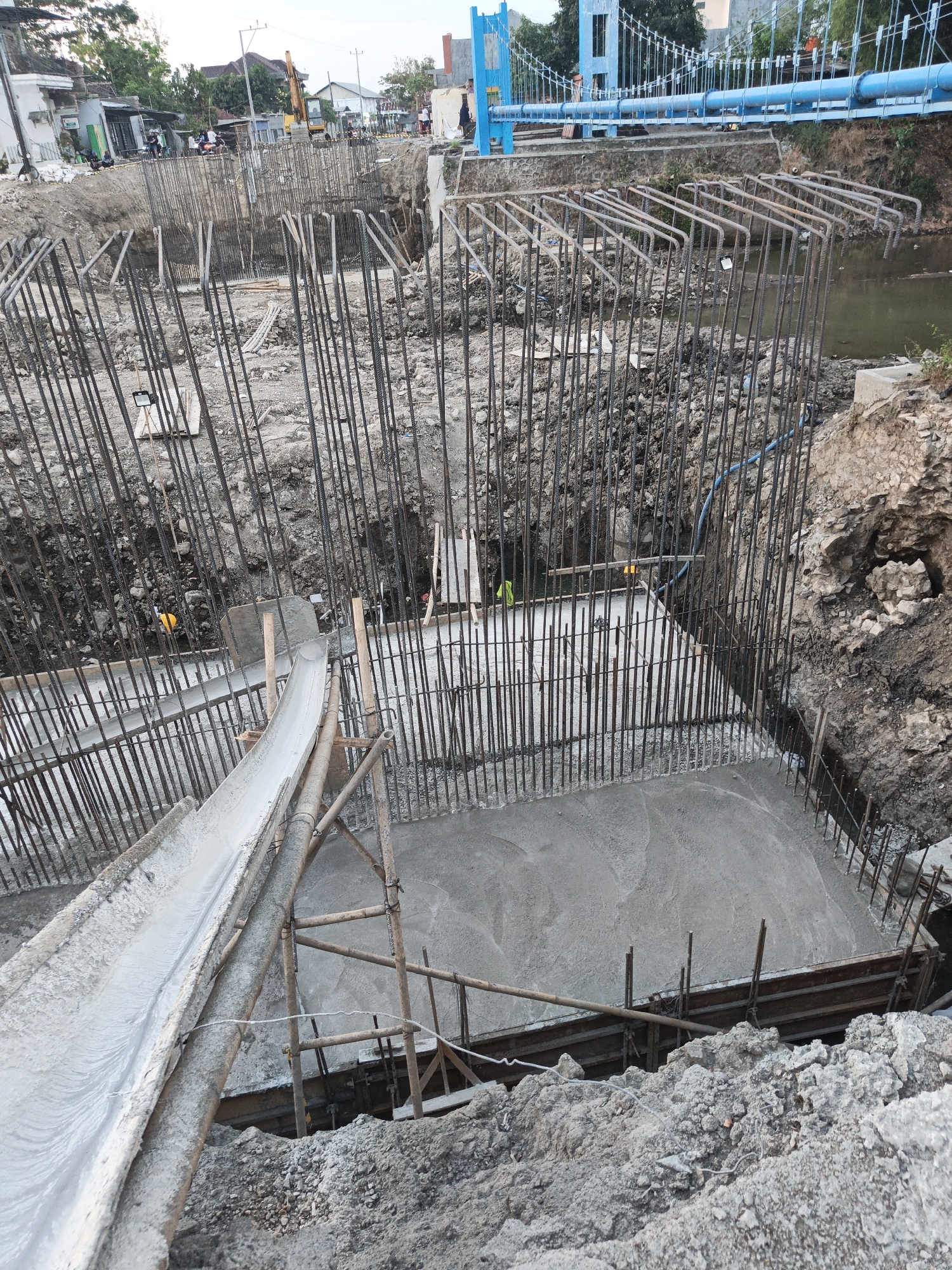 This image depicts a construction site in Planggu, Indonesia, focusing on the foundation work for a bridge. A large, rectangular pit has been excavated, filled with a dense network of vertical steel rebar structures that form the framework for concrete pillars. In the foreground, a concrete chute extends from the upper left, delivering freshly mixed concrete into the base of the pit, where a level layer of cement is being laid. The surrounding area is composed of loose soil, dirt, and rocks, with some scaffolding made of bamboo visible. In the background, a blue suspension bridge is partially visible, suggesting that the construction is related to this existing infrastructure. Several individuals, dressed in casual work attire, are present on the construction site, overseeing the concrete pouring process. The overall scene is characteristic of heavy civil engineering construction.