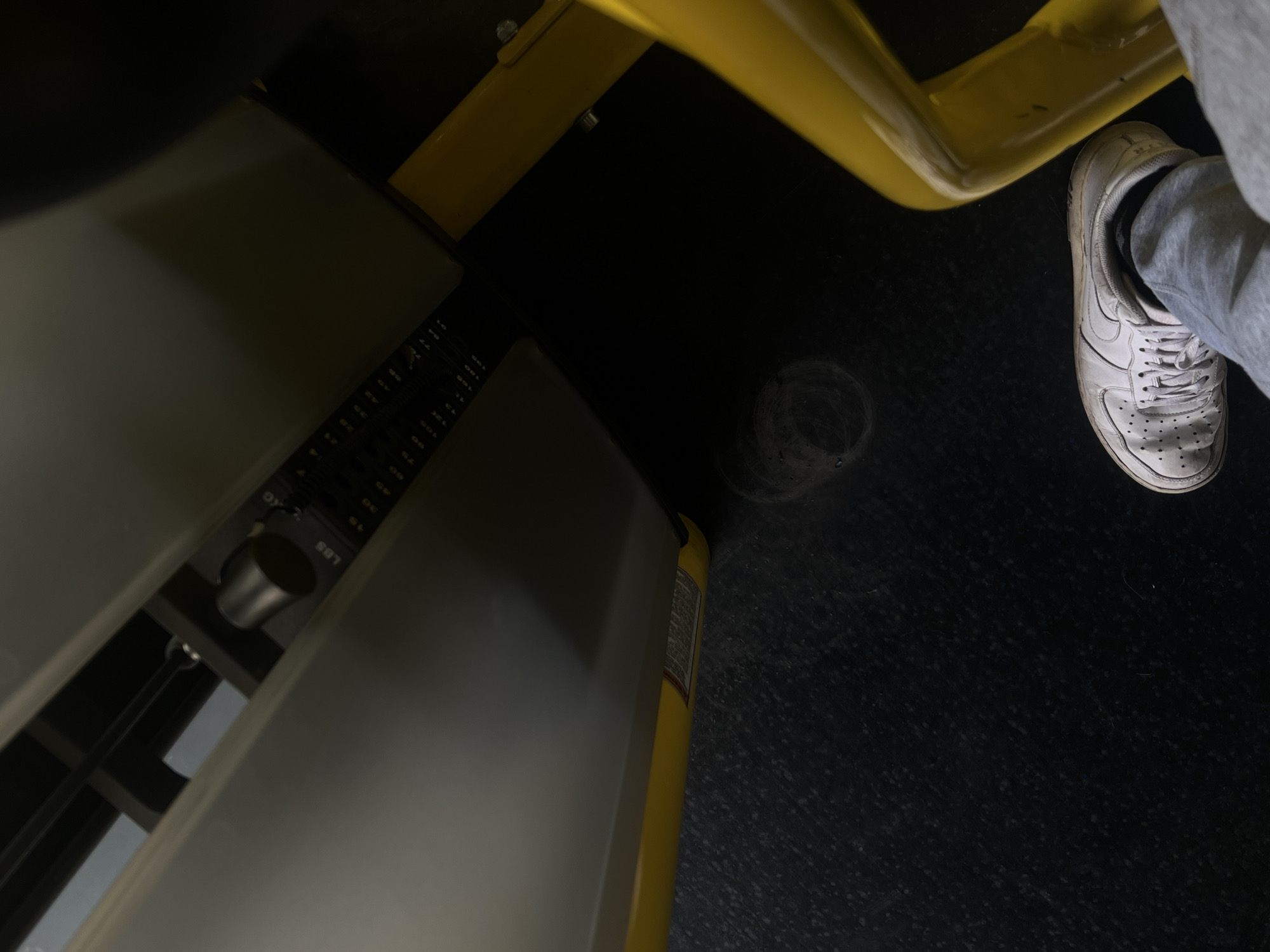 An overhead, close-up shot captures a partial view of a gym setting, focusing on a person's foot and a piece of exercise equipment. The scene is dominated by a dark, speckled floor, likely rubberized, characteristic of a fitness center.
In the upper right quadrant, a person's right foot is visible, clad in a white sneaker (appearing to be Nike Air Force 1s, with the swoosh logo faintly visible) and grey sweatpants. The foot is positioned near a bright yellow metal frame, which could be part of the gym equipment or a bench.
To the left, a large, light-grey weight machine occupies a significant portion of the frame. Part of its mechanism is visible, including a weight selection pin inserted into a stack of weights. Text on this mechanism clearly shows "LBS" (pounds) and "KG" (kilograms), indicating the units of measurement. Visible numbers on the weight stack include "45", "50", "70", "77", "80", "100", and "110", suggesting the weight options.
The lighting is somewhat dim, creating a slightly moody atmosphere, typical of some indoor gym environments. No specific time of day or weather can be determined from the indoor setting. The composition suggests a personal perspective, possibly taken by the person whose foot is shown, capturing their immediate surroundings while engaged in or resting during a workout. There's a faint circular mark on the floor near the yellow frame. A small, illegible label is visible on the side of the grey machine.