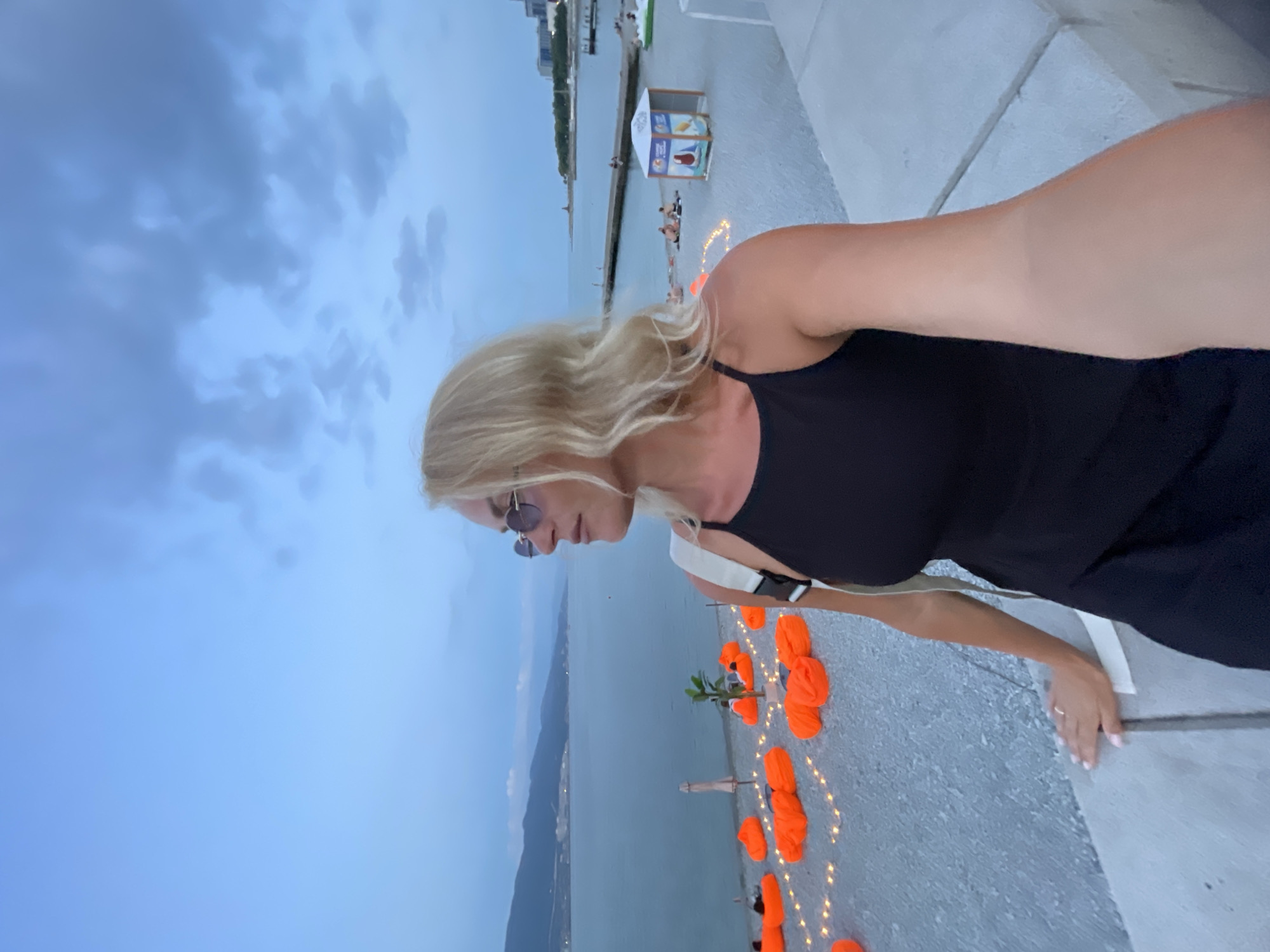 The image shows a woman with blonde hair and sunglasses taking a selfie on a beach in Novorossiysk, Russia. She is wearing a black dress and a white strap across her shoulder. In the background, there is a calm sea, a beach with orange beanbag chairs and string lights, and a pier. The sky is cloudy and appears to be twilight. There are a few other people on the beach in the distance, and some buildings are visible along the coastline.