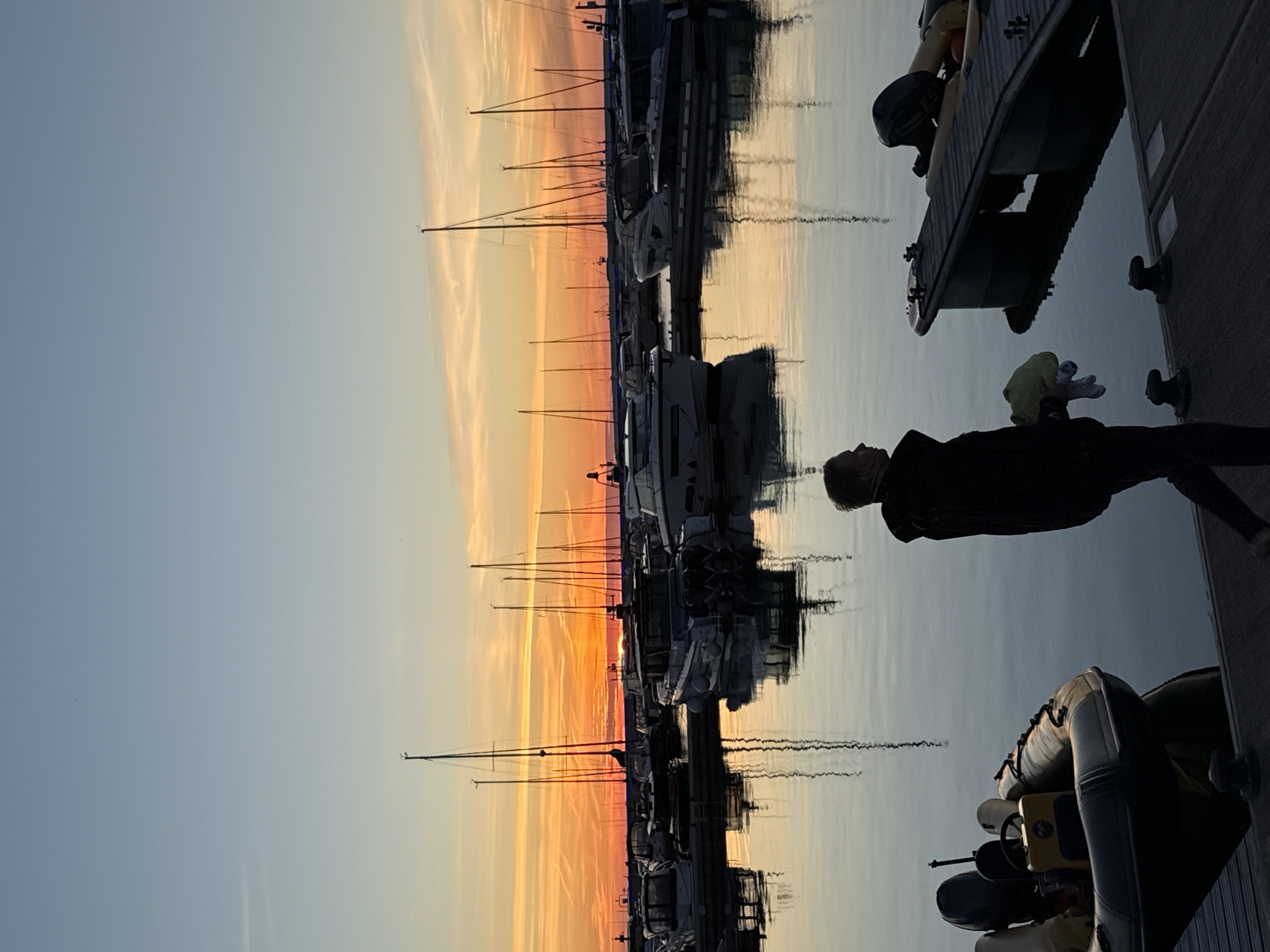 An outdoor, eye-level shot captures a marina at dusk, likely in Tallinn, Estonia, given the context. The sky is a gradient of soft blue to warm orange hues, with streaks of light clouds. A calm body of water reflects the sky and the silhouettes of numerous boats docked in the marina. The boats are of various sizes, including sailboats with tall masts and motorboats. In the foreground, a person, viewed from the side and back, walks along a wooden pier. They are wearing dark clothing and carrying a light-colored object in their right arm. The pier has dark bollards. To the left, a small inflatable boat with an outboard motor is docked. The scene is tranquil, with the sunset casting a soft glow over the water and the boats.