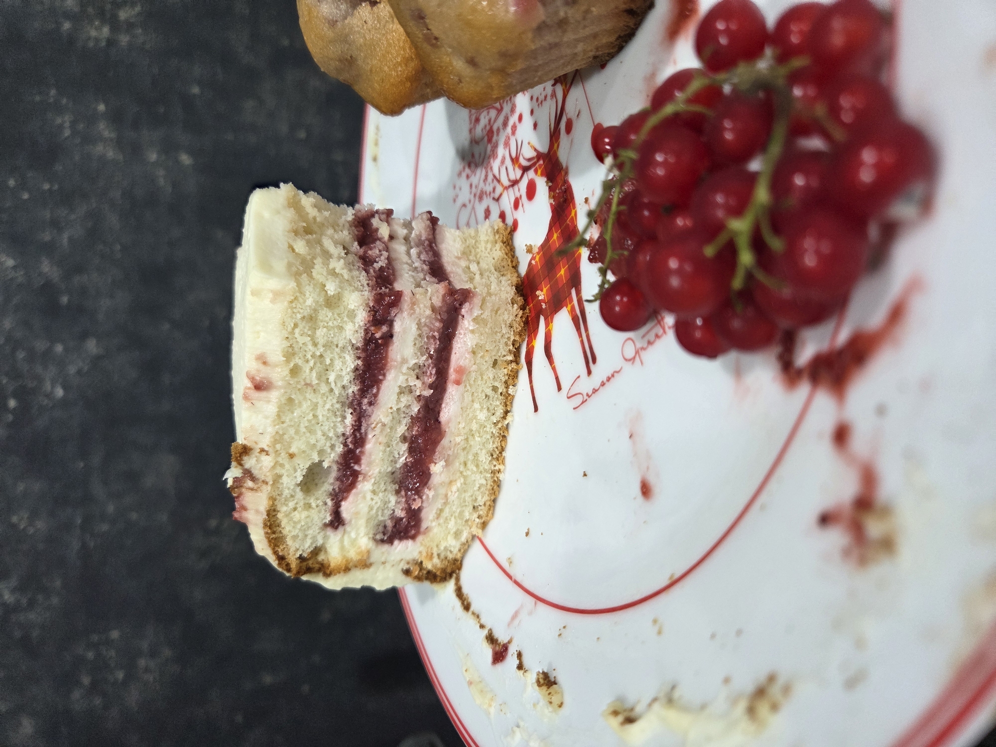 A slice of layered cake with white frosting and red jam filling sits on a white plate with a red rim. Next to the cake are two muffins and a sprig of red currants. The plate has a red deer design. The background is a dark, textured surface.