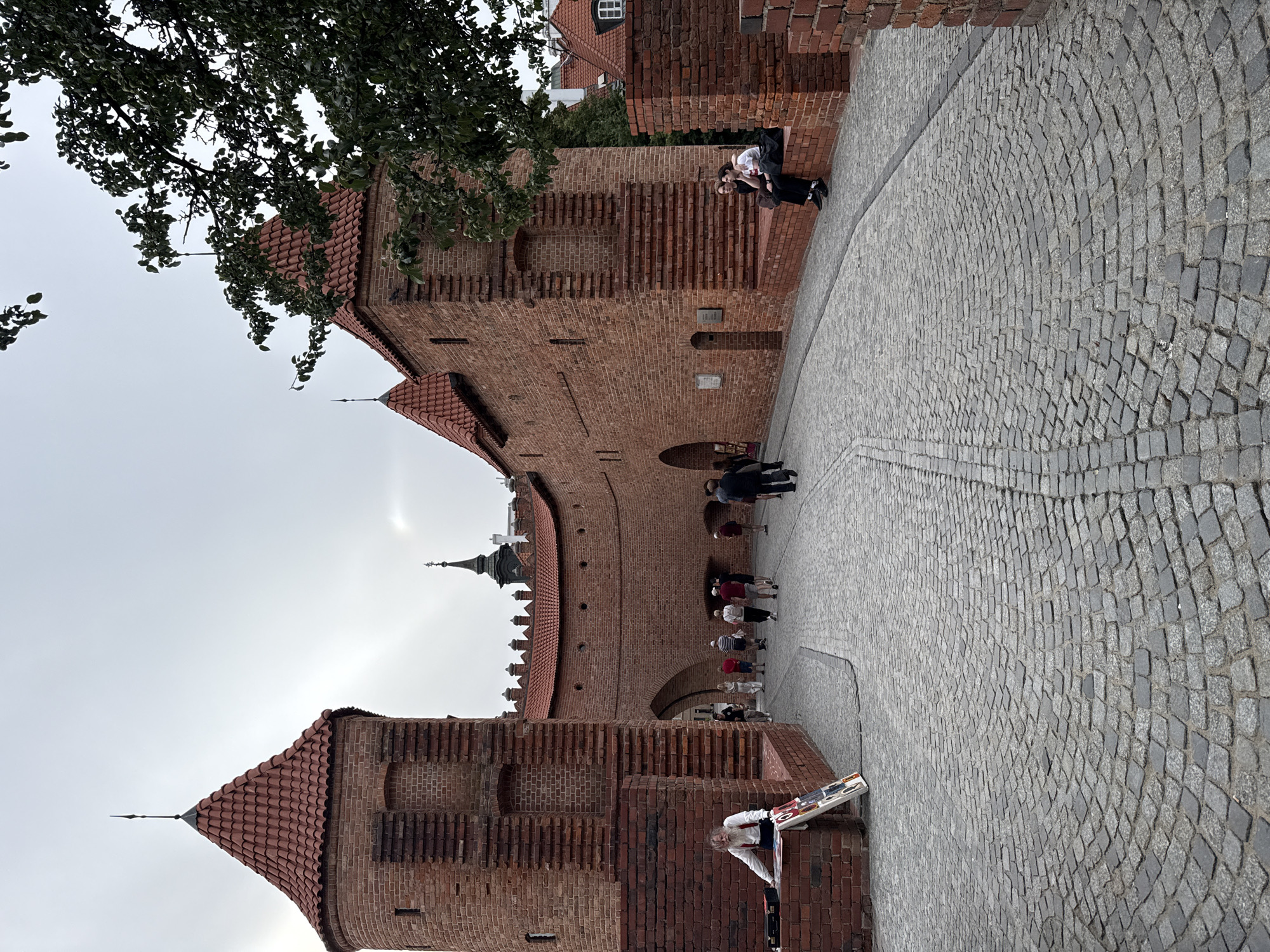 In this image, two young people are sitting on a brick ledge, with the taller of the two resting their arm on the other's shoulder. The background is dominated by a large, ancient brick structure with a red-tiled roof and a cobblestone pathway in front of it. Several other people are walking along the pathway and through an arched entrance of the brick structure. A street vendor with a cart is also visible in the foreground, displaying colorful items. The sky is overcast, suggesting a cloudy day. The scene appears to be a historic location, likely a castle or fortified wall, given the architecture. The overall mood is casual and observational, capturing a moment of daily life in a historical setting. The location is identified as Warsaw, Poland, which aligns with the architectural style. No discernible text is visible in the image.