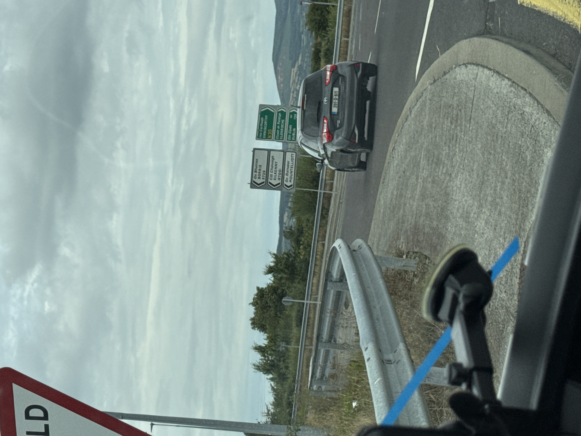 The image shows a grey Toyota Yaris driving on a road. The car has a license plate that reads "201-D-611". In the background, there are several road signs. One sign indicates directions to "Borrris", "KILKENNY", and "MOUNTIELLOT". Another sign points towards "ENNISCORTHY", "CLONROCHE", and "DUBLIN". The sky is overcast and there are hills in the distance. The location appears to be in or near New Ross Municipal District, Ireland, as indicated by the road signs.