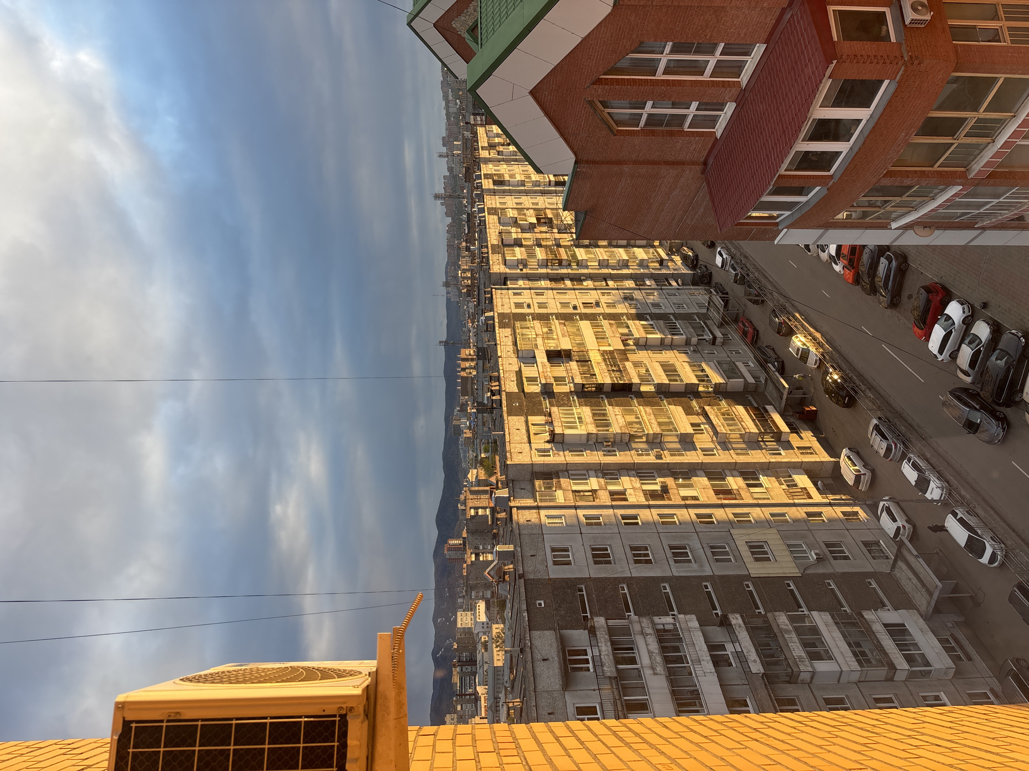 The image is a high-angle, almost aerial view of an urban landscape, presumably taken from a tall building in Krasnoyarsk, Russia. The scene is dominated by numerous multi-story apartment buildings, many equipped with air conditioning units on their facades. The buildings vary in architectural style and condition, some appearing newer and better maintained than others. A street runs alongside the buildings, lined with parked cars of various colors. In the background, a distant mountain range can be seen under a sky with patchy clouds. The sun is low, casting long shadows across the buildings and street, indicating either early morning or late afternoon. The overall impression is that of a densely populated residential area in a city setting.