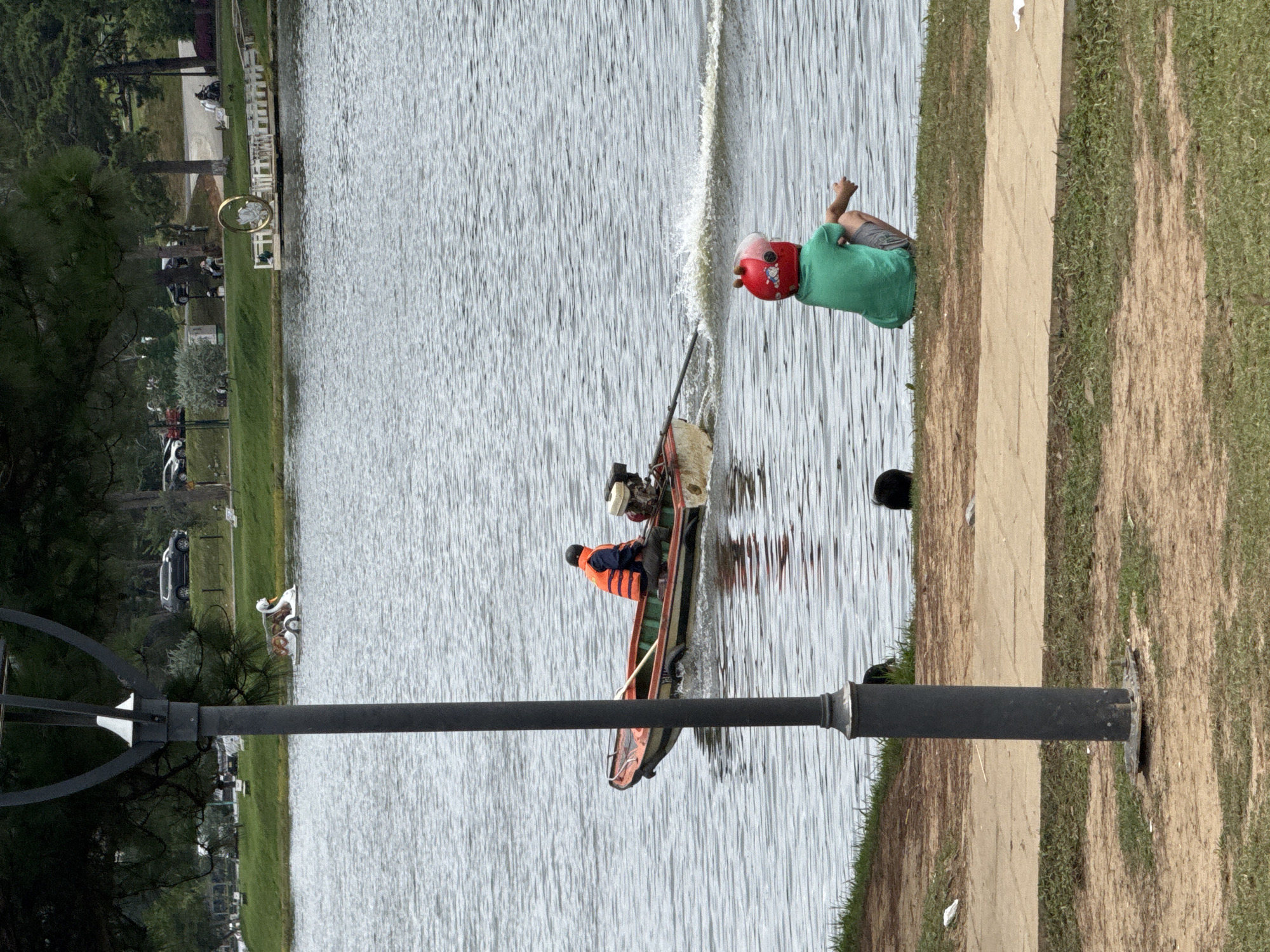 This is a photograph of Xuan Huong Lake in Da Lat, Vietnam. In the foreground, a child wearing a green t-shirt and a red helmet with a pink visor sits on the grassy bank, facing the water. In the middle ground, a small motorboat with two occupants speeds across the lake, creating a wake. The person in the back of the boat wears an orange life vest. In the background, a swan-shaped pedal boat is visible on the water, as well as trees, a road with cars, and a large golden ring structure on a pier. A lamppost stands prominently in the left foreground. The overall scene depicts a recreational area with various activities on the lake.