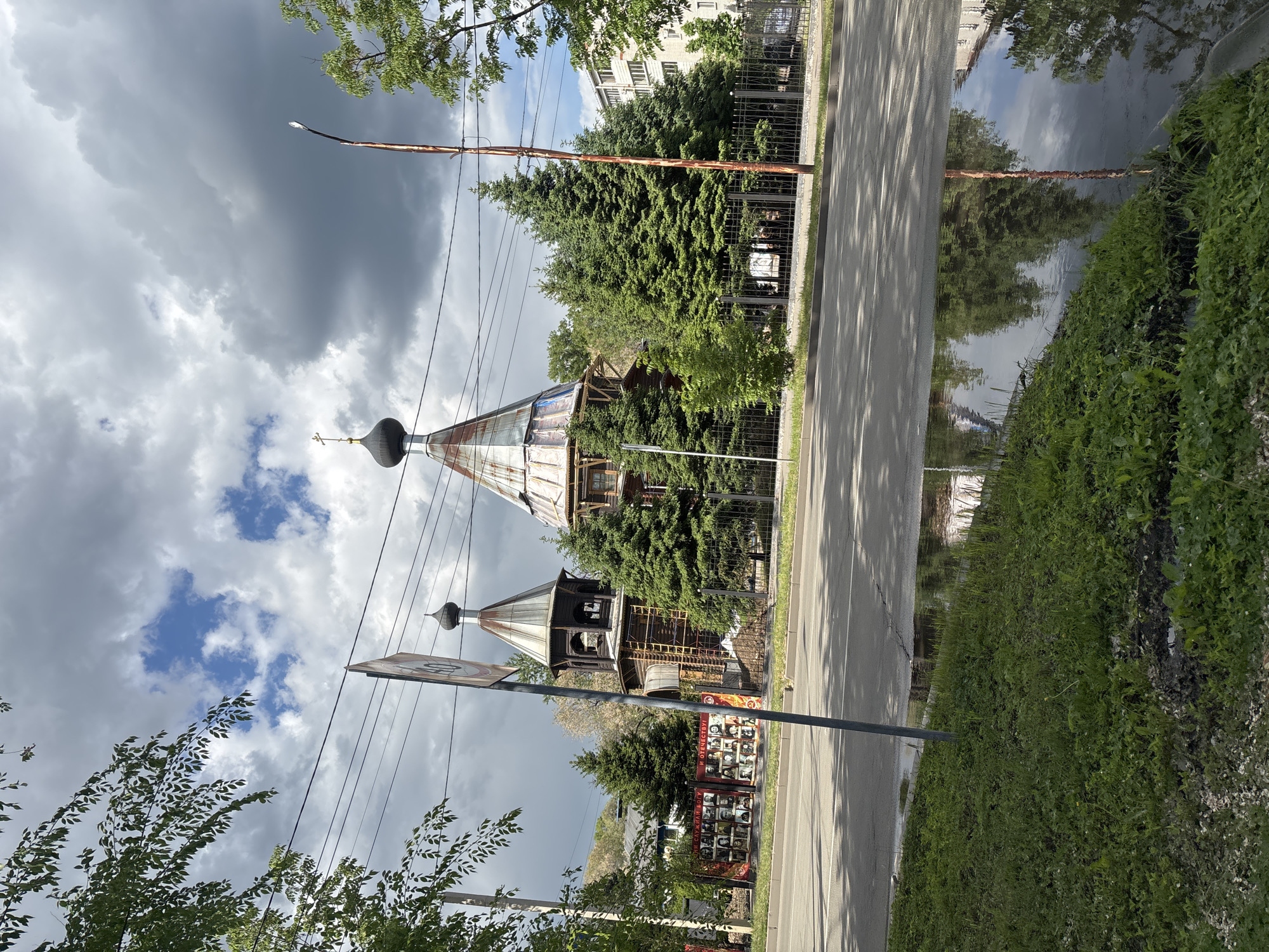 This outdoor photograph captures a street scene featuring two wooden structures resembling Orthodox churches or chapels, set against a backdrop of trees and an overcast sky. The image appears to be taken during the daytime, with varying light suggesting partly cloudy weather.
In the center-left, two wooden buildings with distinctive onion domes and crosses are visible. The larger, taller structure seems to be under construction or renovation, with scaffolding-like elements and unfinished cladding on its upper section. Both domes are dark, possibly black, with small golden crosses on top. Dense coniferous trees partially obscure the buildings.
To the right of the structures, a paved road or pathway runs parallel to a metal fence, separating it from the property. Further right, a lush green grassy area extends, bordering a large reflective surface that appears to be water, possibly a wide puddle or a small body of water, mirroring the sky, trees, and a utility pole. The pole, which is reddish-brown and supports a street lamp and multiple wires, stands prominently near the road.
In the background, behind the trees, a light-colored multi-story building is visible. In the foreground, on the left, parts of leafy deciduous trees frame the scene. The sky above is largely covered by dark, heavy clouds, with glimpses of brighter blue sky and white clouds breaking through.
A circular traffic sign with the number "50" is partially visible in the lower-left corner. Behind the smaller wooden structure, a rectangular sign displays multiple portrait photographs and some Cyrillic text, including "И ОТЕЧЕСТВУ!" (And Fatherland!) and what might be "ДРУГАЯ ИМЯ БОГУ" (Another Name for God) or a similar phrase, suggesting a memorial or tribute. No people are visible, and no specific activity is taking place. The overall impression is one of a quiet, somewhat overgrown urban or suburban area with traditional religious architecture.