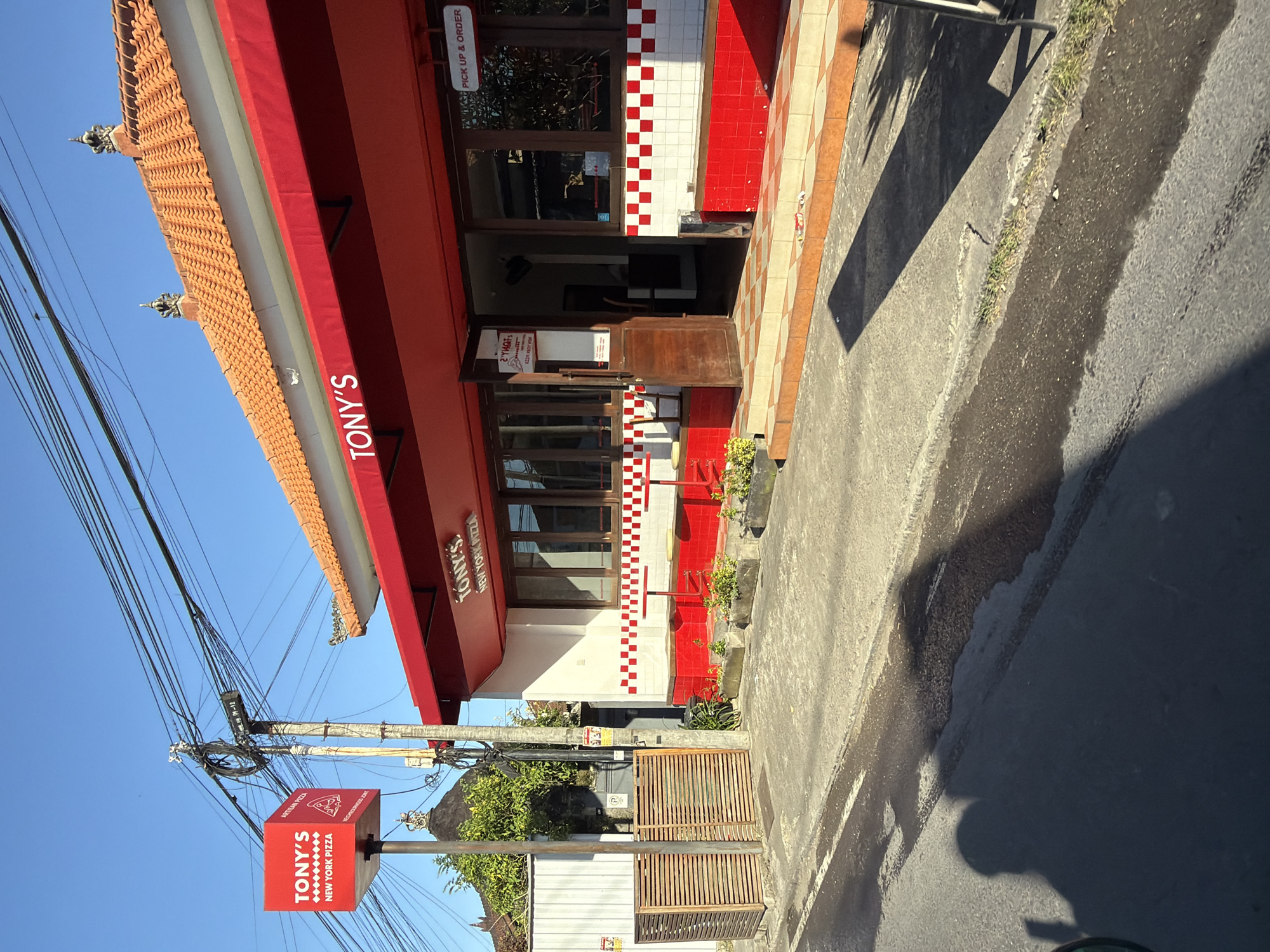 Here is a description of the image:
The image shows the exterior of "Tony's New York Pizza" located in what appears to be Mengwi, Indonesia. The building has a red and white color scheme, with red awnings over windows and a traditional-style brown tiled roof with decorative elements at the corners. The front of the building has alternating red and white tiles arranged in a checkerboard pattern near the entrance. A sign at the street level clearly displays "TONY'S NEW YORK PIZZA" on a red background. Overhead, a smaller sign indicates "PICK UP & ORDER".
The street area is paved and shows some water or dampness on the surface. Above the building, there is a dense web of utility wires against a clear blue sky. The building is surrounded by vegetation, including trees and small plants.