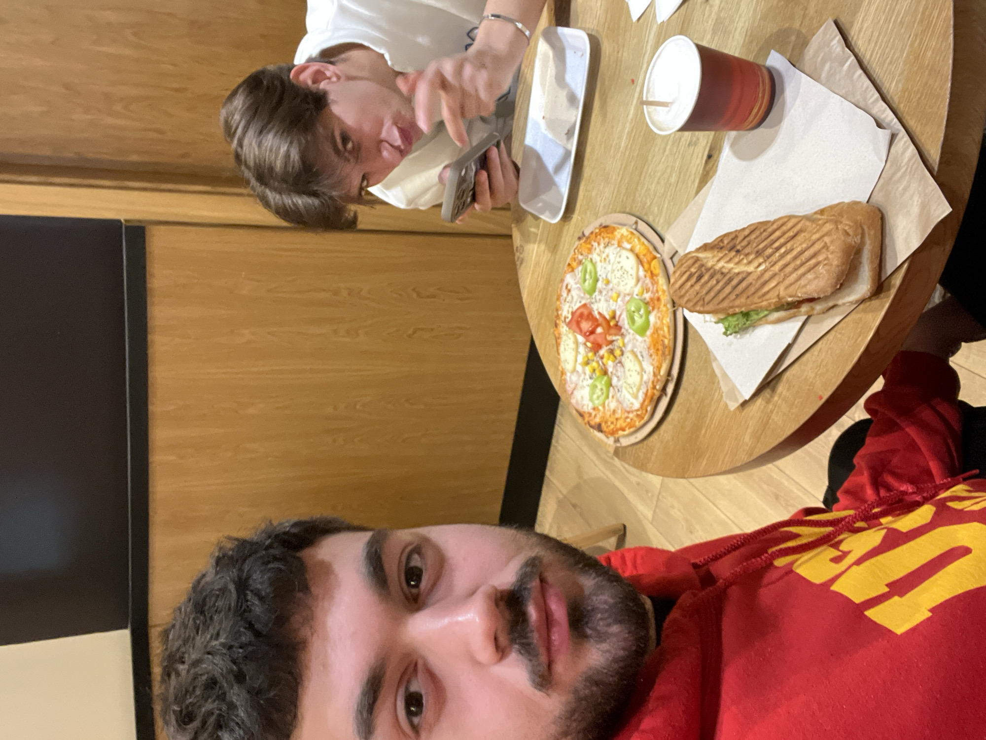 The image is a selfie taken at a table with food and two people. The person in the foreground wears a red hoodie with yellow lettering. Behind them, a round wooden table holds a pizza, a sandwich, and a drink. The pizza is topped with mozzarella, corn, tomatoes, and green peppers. The sandwich is grilled and contains lettuce. The drink is in a red paper cup with a white lid and a straw. A second person in a white t-shirt is visible in the background, holding a phone. The location is likely Sofia, Bulgaria, but this can not be confirmed from the image. The environment appears to be indoors, possibly a restaurant or cafe, with wood paneling on the wall.