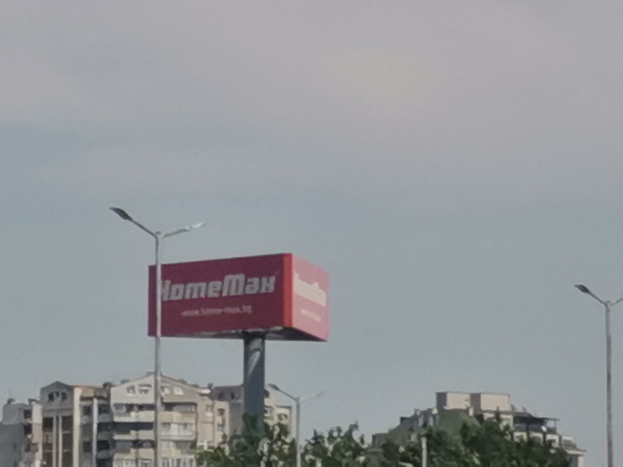 This image captures a daytime scene under an overcast or hazy light grey-blue sky, likely in Plovdiv, Bulgaria, as indicated by the "www.home-max.bg" URL on the prominent billboard. The central subject is a large, rectangular red billboard displaying "HomeMax" in white Cyrillic script and "www.home-max.bg" in smaller white text below it, supported by a thick grey pole.
In the foreground, a tall, slender street lamp with a single light fixture extends upwards on the left, partially overlapping the billboard pole. Another similar street lamp is visible on the far right edge of the image.
Below the billboard and street lamps, a row of multi-story buildings, predominantly light-colored and featuring balconies and varied architectural details, stretches across the mid-ground. Green foliage, likely trees or bushes, partially obscures the lower sections of these buildings and the base of the billboard pole. No individuals or specific actions are discernible within the frame.